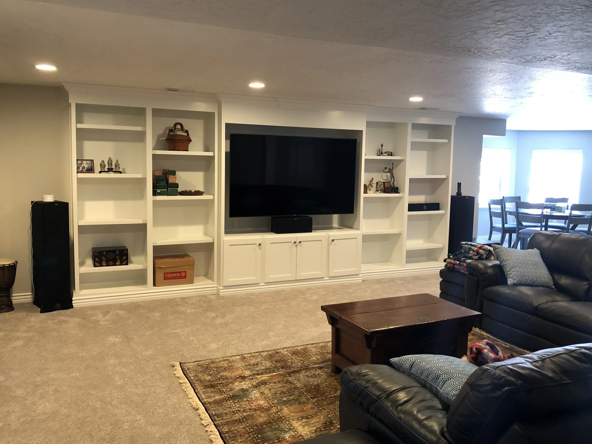 Living room with beige carpet, white built-in shelves, large flat-screen TV, black speakers, brown wooden coffee table, black leather sofas, and a dining area with a table and chairs in the background.