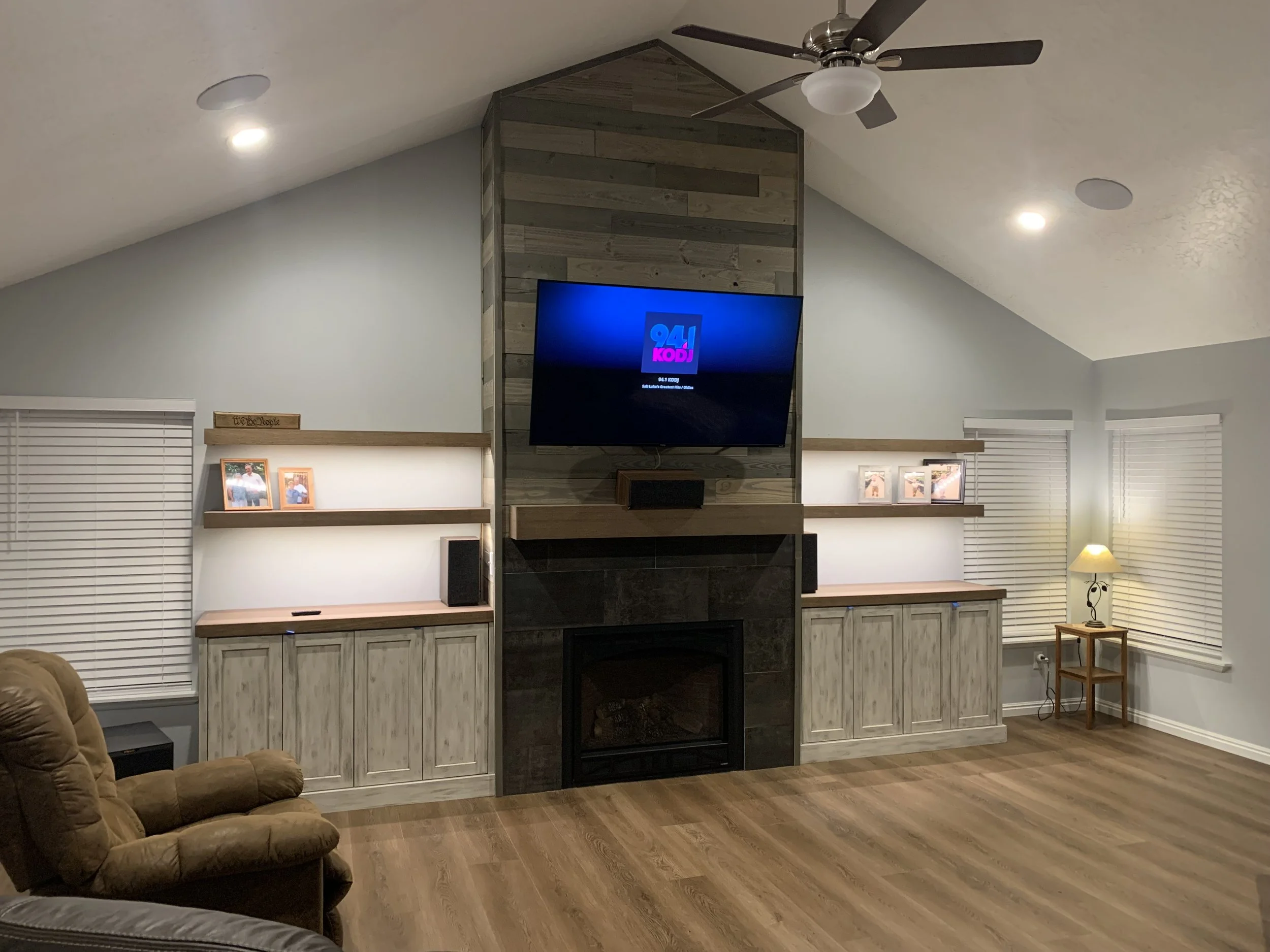 Living room with a log cabin style fireplace, a large flat-screen TV mounted above it, and built-in cabinets on either side. The room has wooden flooring, a recliner chair on the left, white blinds on the windows, a small side table with a lamp on th