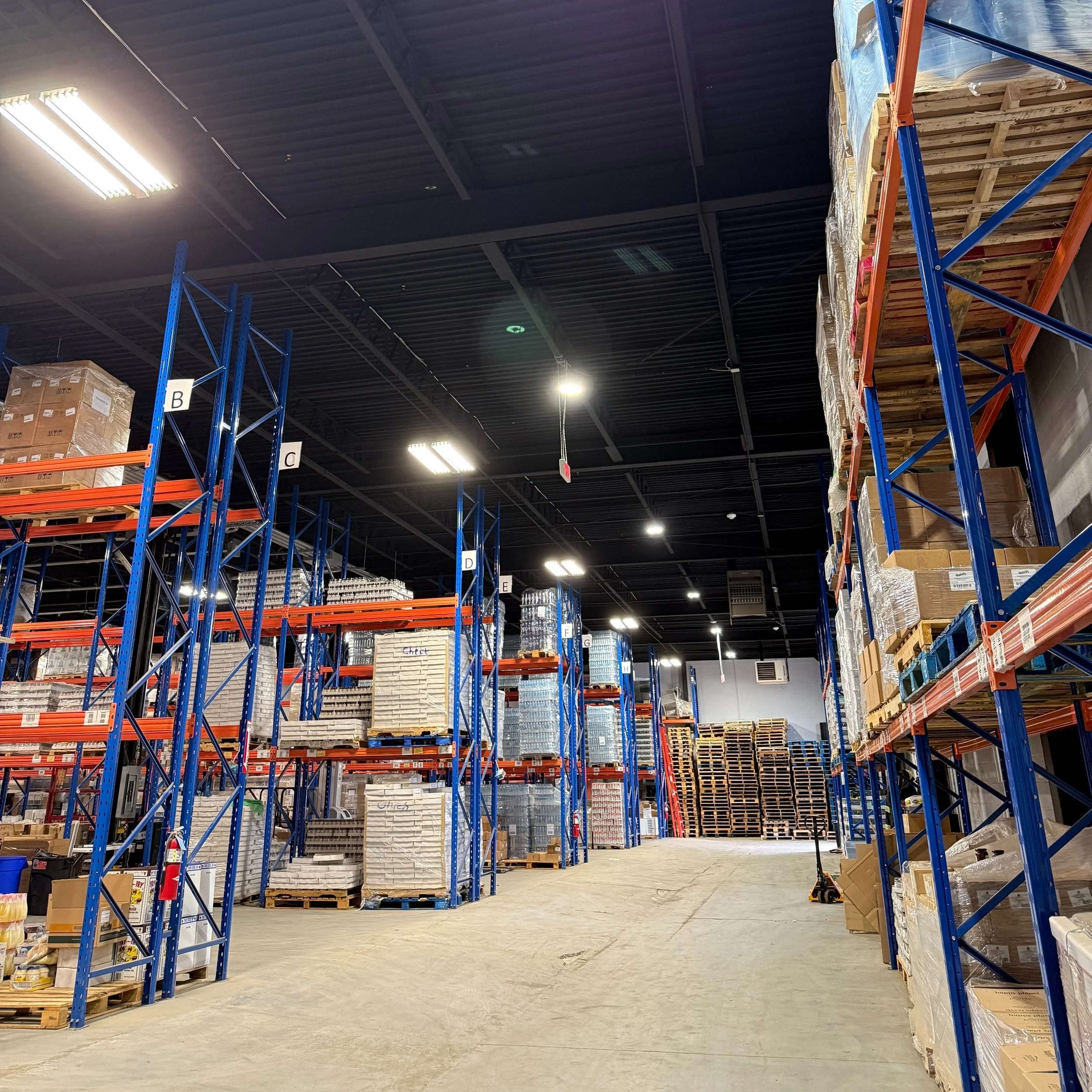 Inside a warehouse with empty metal shelving units, some filled with boxes, pallets, and supplies, against a dark ceiling with overhead lighting.