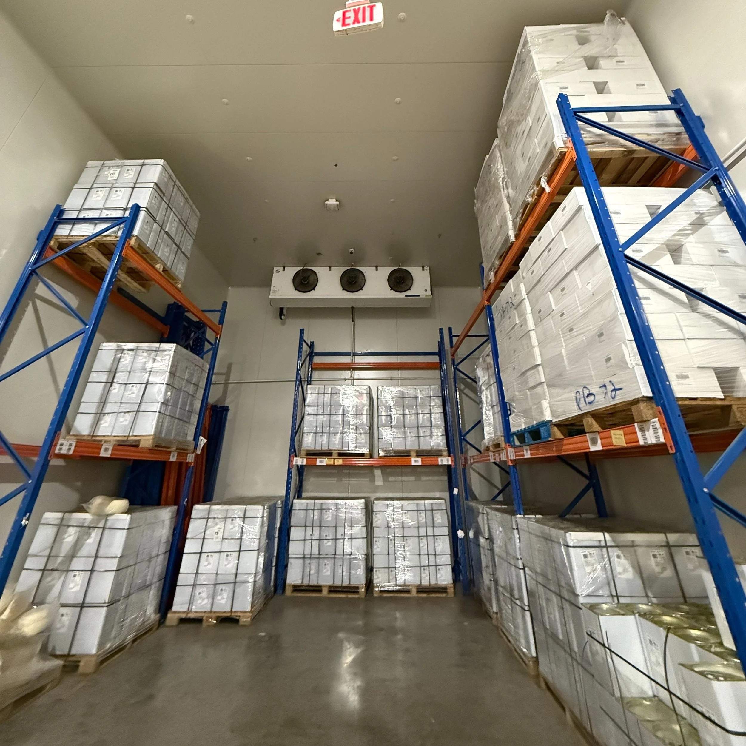 Storage room with metal shelving units holding boxes wrapped in plastic, with an overhead exhaust system in the background.