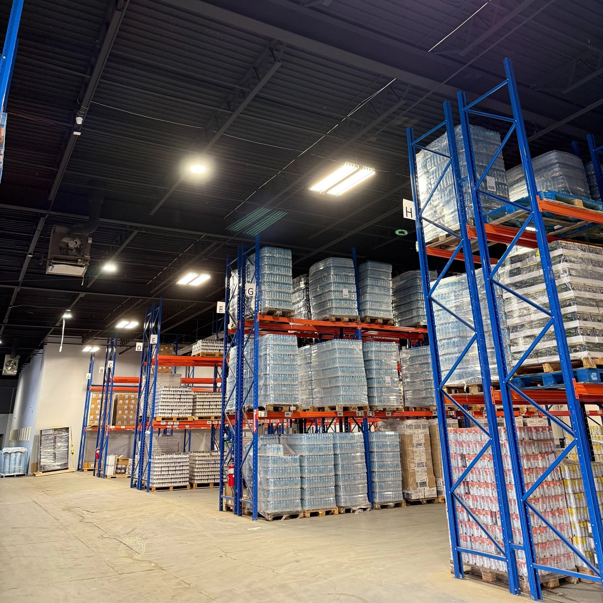 Warehouse with tall blue and orange metal shelves filled with stacked pallets of bottled water and other goods.