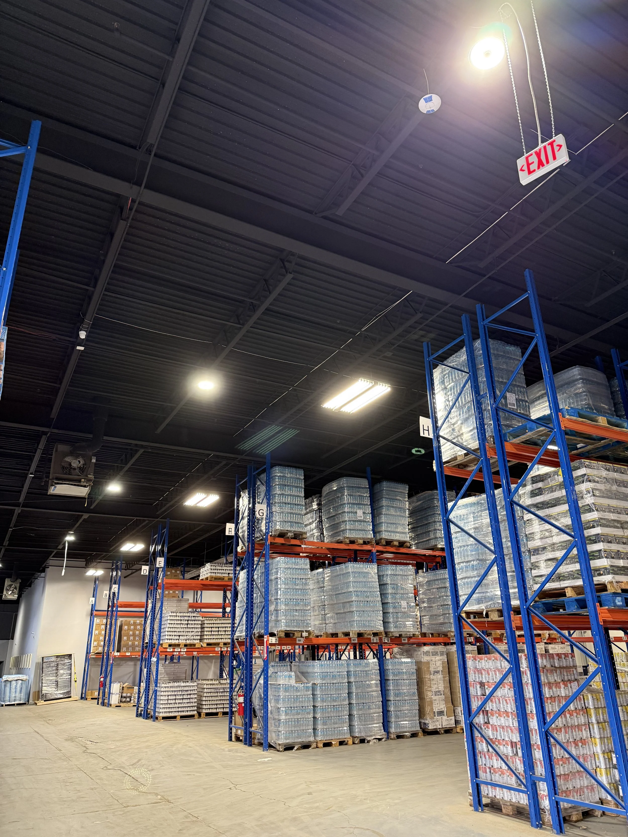 A warehouse with tall blue and orange shelving units filled with stacked pallets of goods, under a black ceiling with fluorescent lighting and an illuminated exit sign.