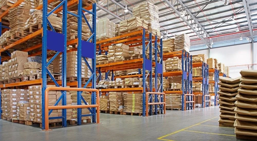 Warehouse with multiple tall orange and blue storage racks filled with stacked brown paper packages and boxes, some wrapped in plastic, and large stacks of flattened brown cardboard boxes on the floor.