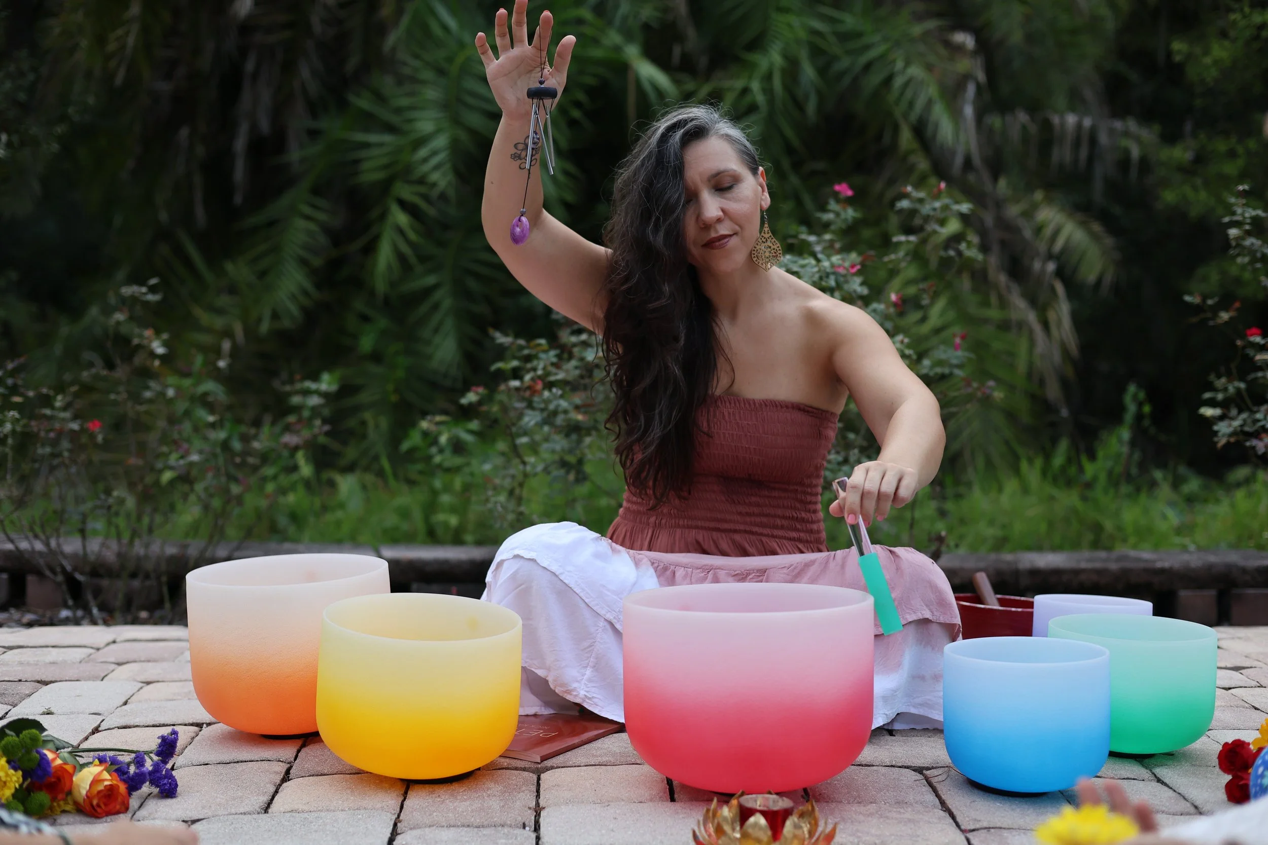 Woman playing crystal singing bowls outdoors with lush greenery in the background.