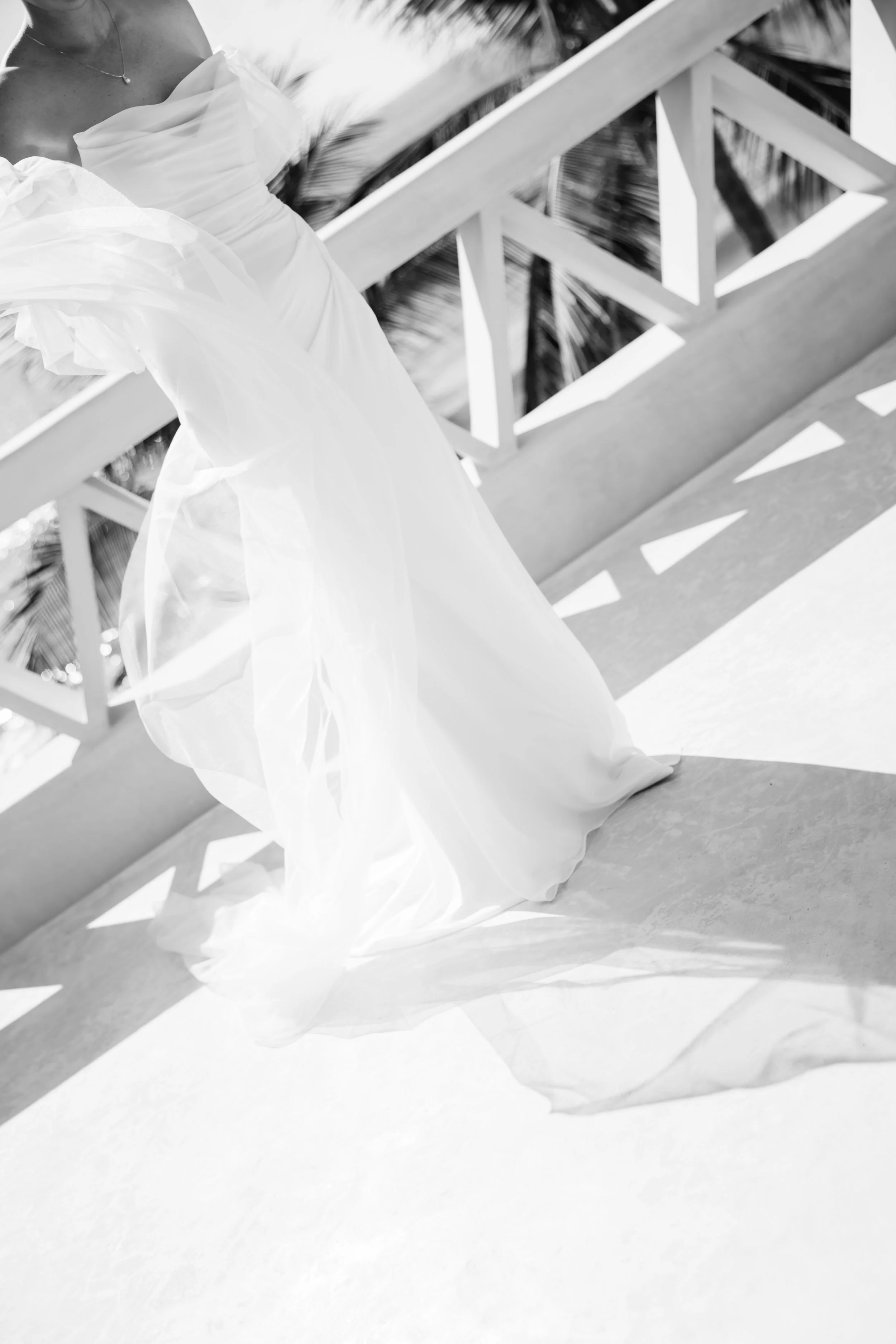 Close-up of a person wearing a flowing white dress on a balcony with a wooden railing and palm trees in the background, in black and white.