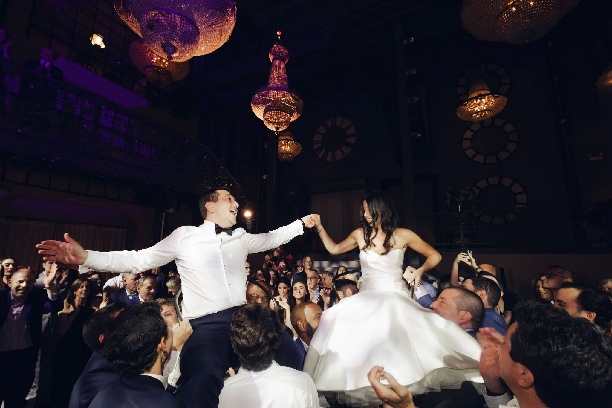 A wedding celebration with the bride and groom dancing on chairs held by guests in a grand reception hall with chandeliers.