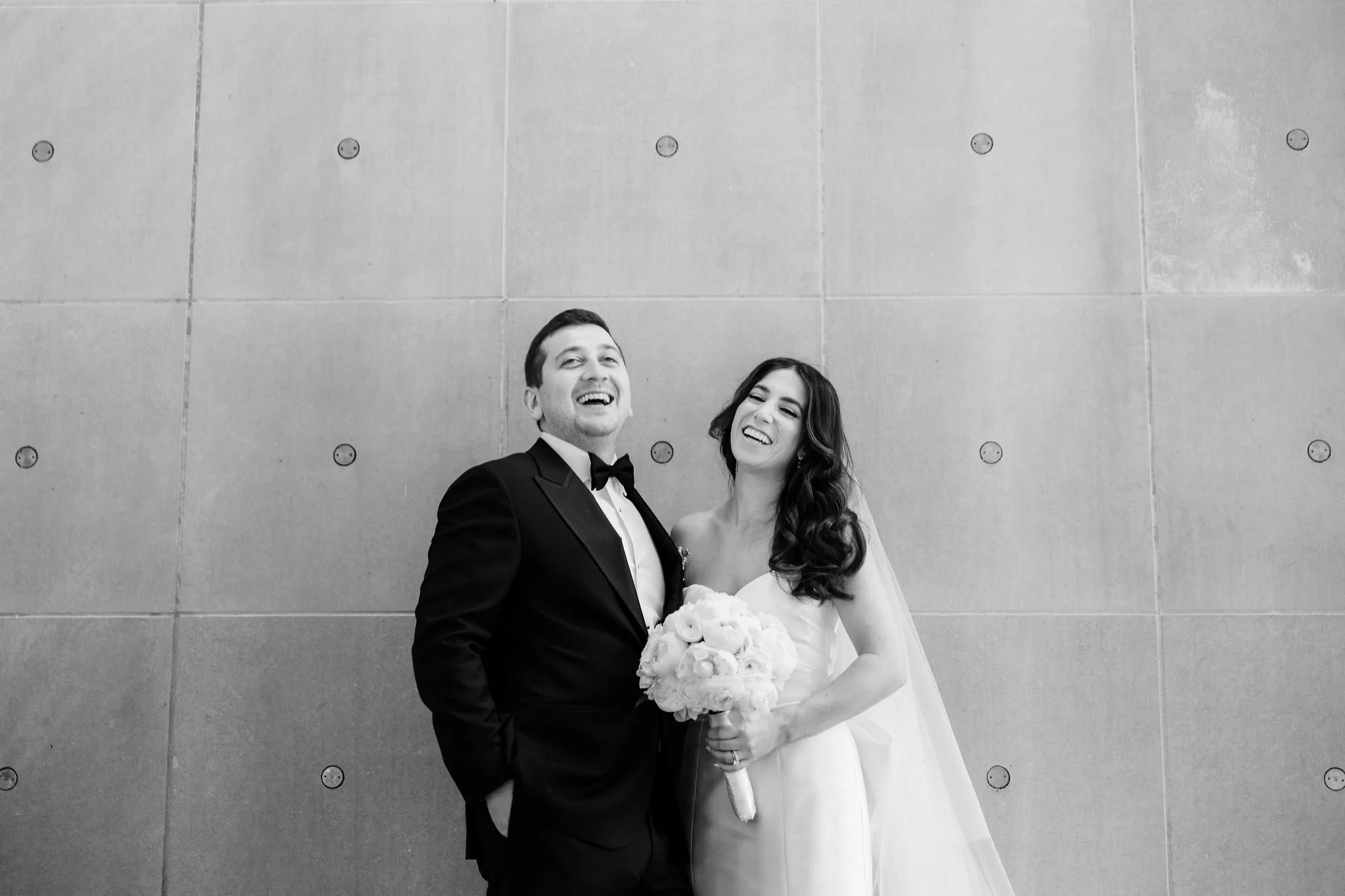 Black and white photograph of a happy bride and groom on their wedding day, standing in front of a textured concrete wall. The groom is wearing a tuxedo with a bow tie, and the bride is in a strapless wedding gown holding a bouquet of flowers. Both a
