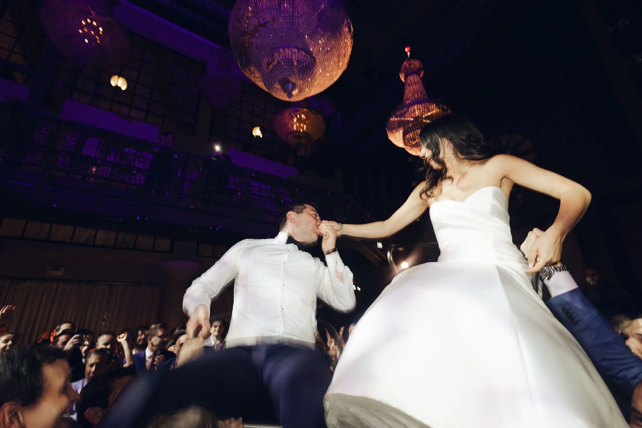 A bride in a white wedding gown and a groom in a tuxedo are dancing at a wedding reception, with guests watching in the background and elegant chandeliers hanging from the ceiling.