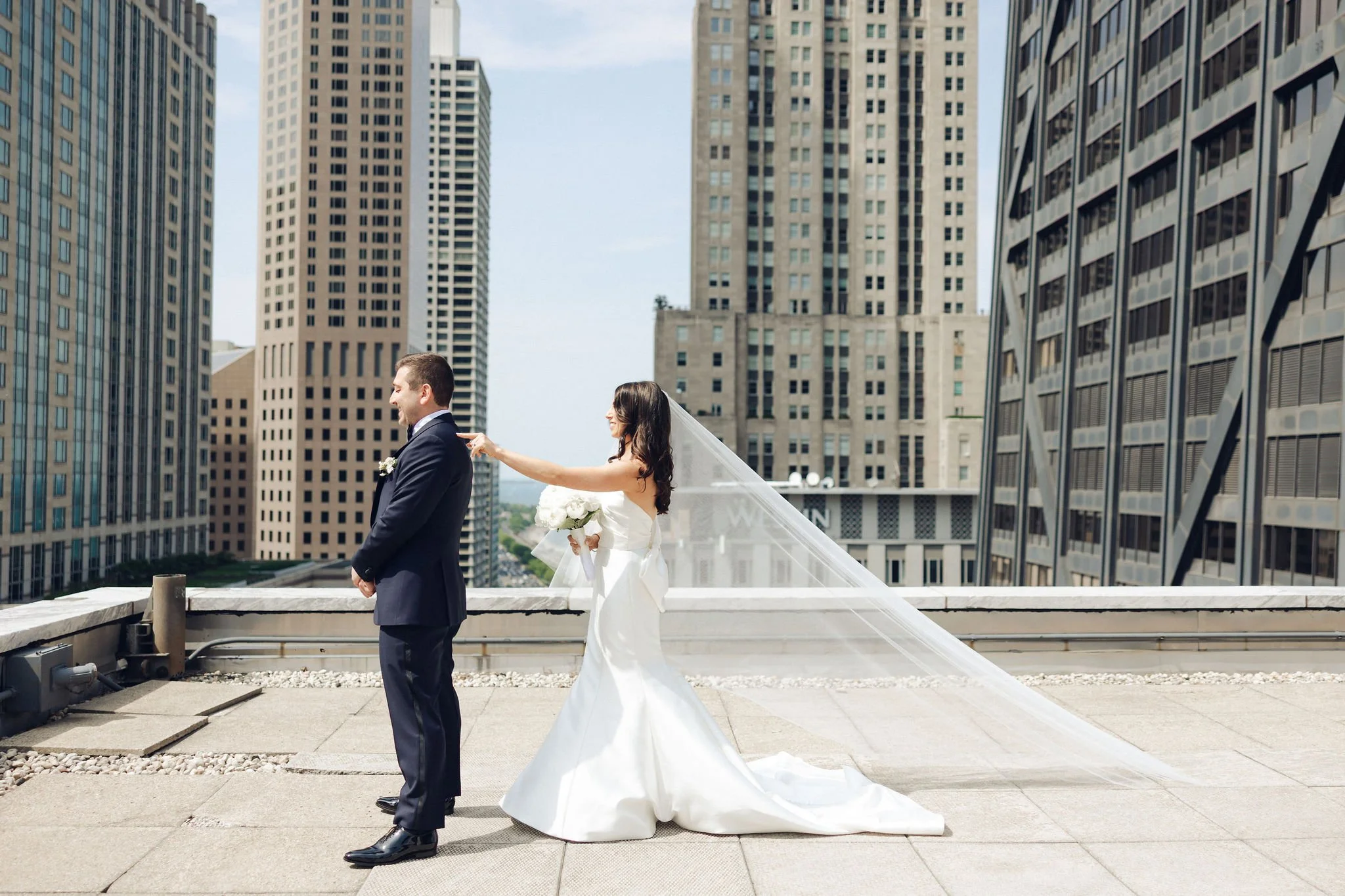 Bride in a white wedding dress playfully pulls the groom's tie on a rooftop with city skyscrapers in the background.
