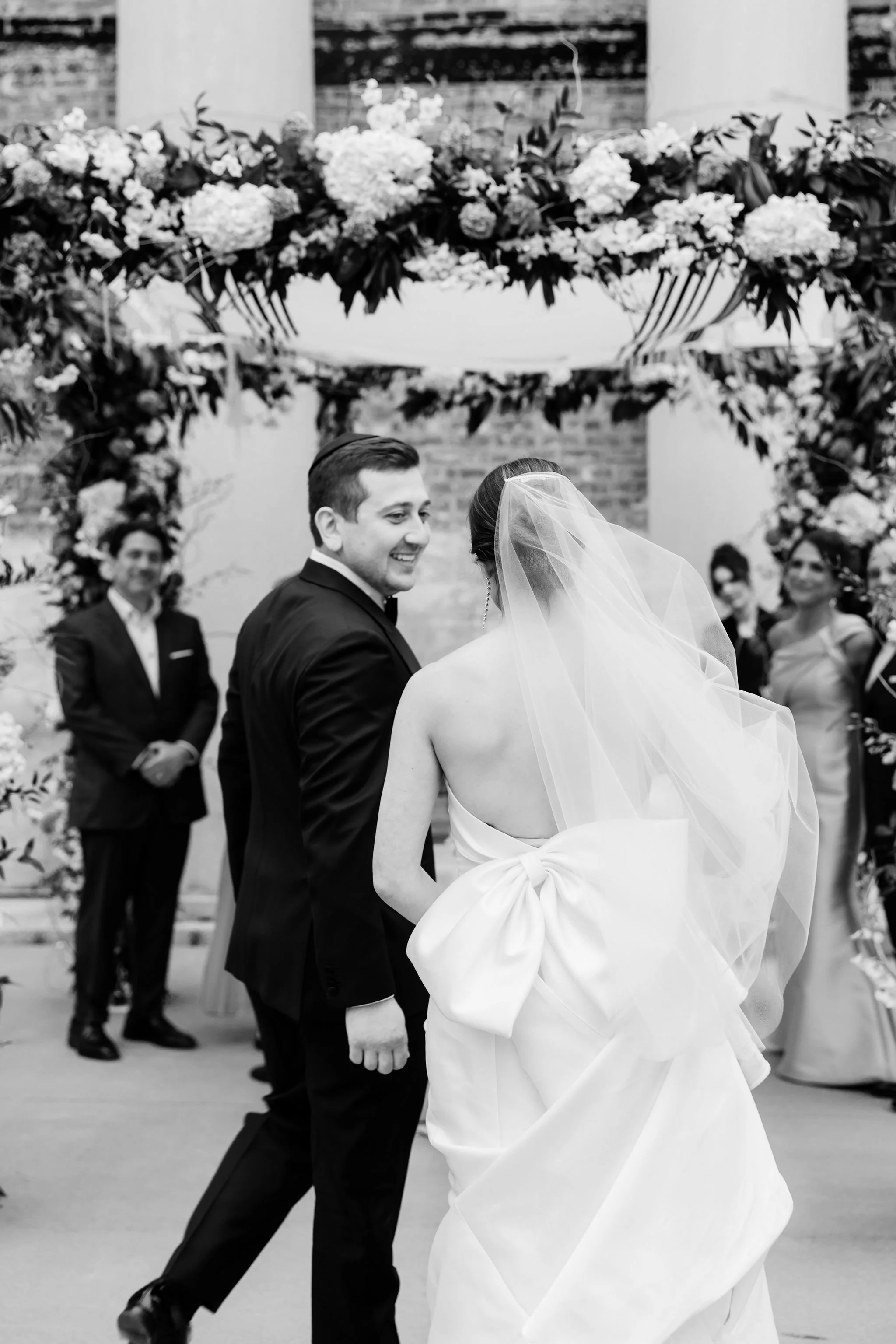 Black and white photo of a wedding ceremony with a groom smiling at the bride, who is wearing a strapless wedding dress with a large bow and veil, under a floral arch with guests in the background.