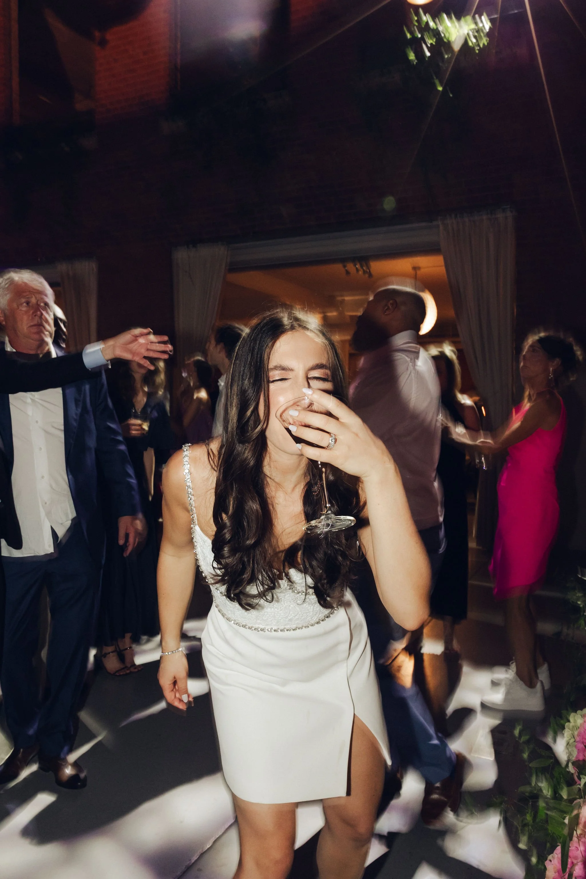 A woman with long dark hair in a white dress is laughing and covering her mouth with her hand while holding a glass of champagne at a lively party or celebration.