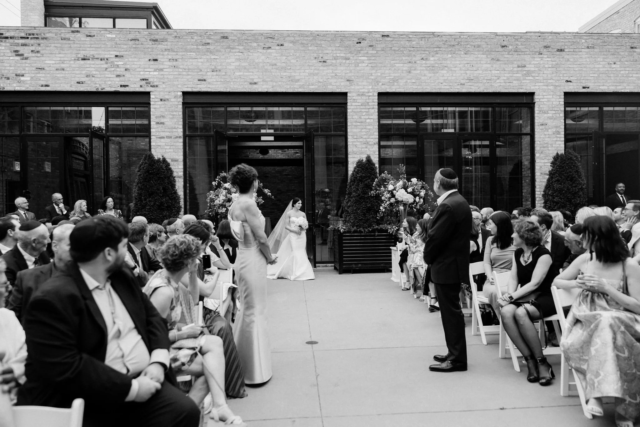 A black and white photo of a wedding ceremony outdoors with guests seated on either side, a bride in a white gown holding a bouquet, and a groom in a dark suit standing in front of her, with a large brick building in the background.