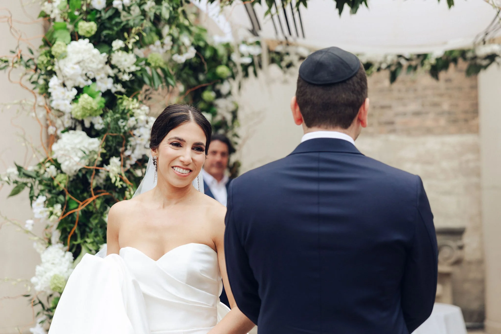 A bride and groom exchange vows during a wedding ceremony outdoors, with a floral arch in the background.