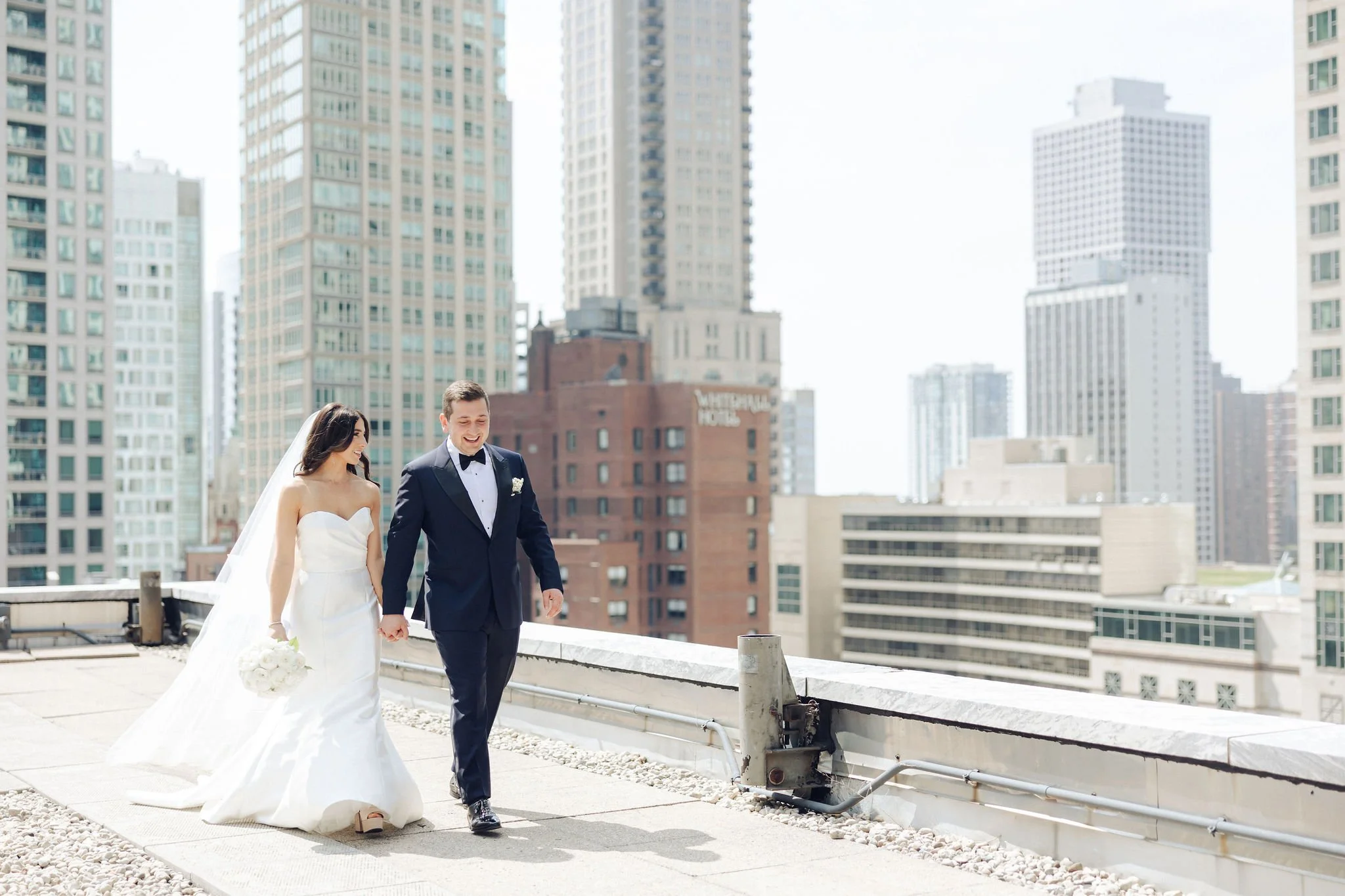 A bride in a white wedding gown holding a bouquet walking with a groom in a dark tuxedo on a rooftop with a city skyline in the background.