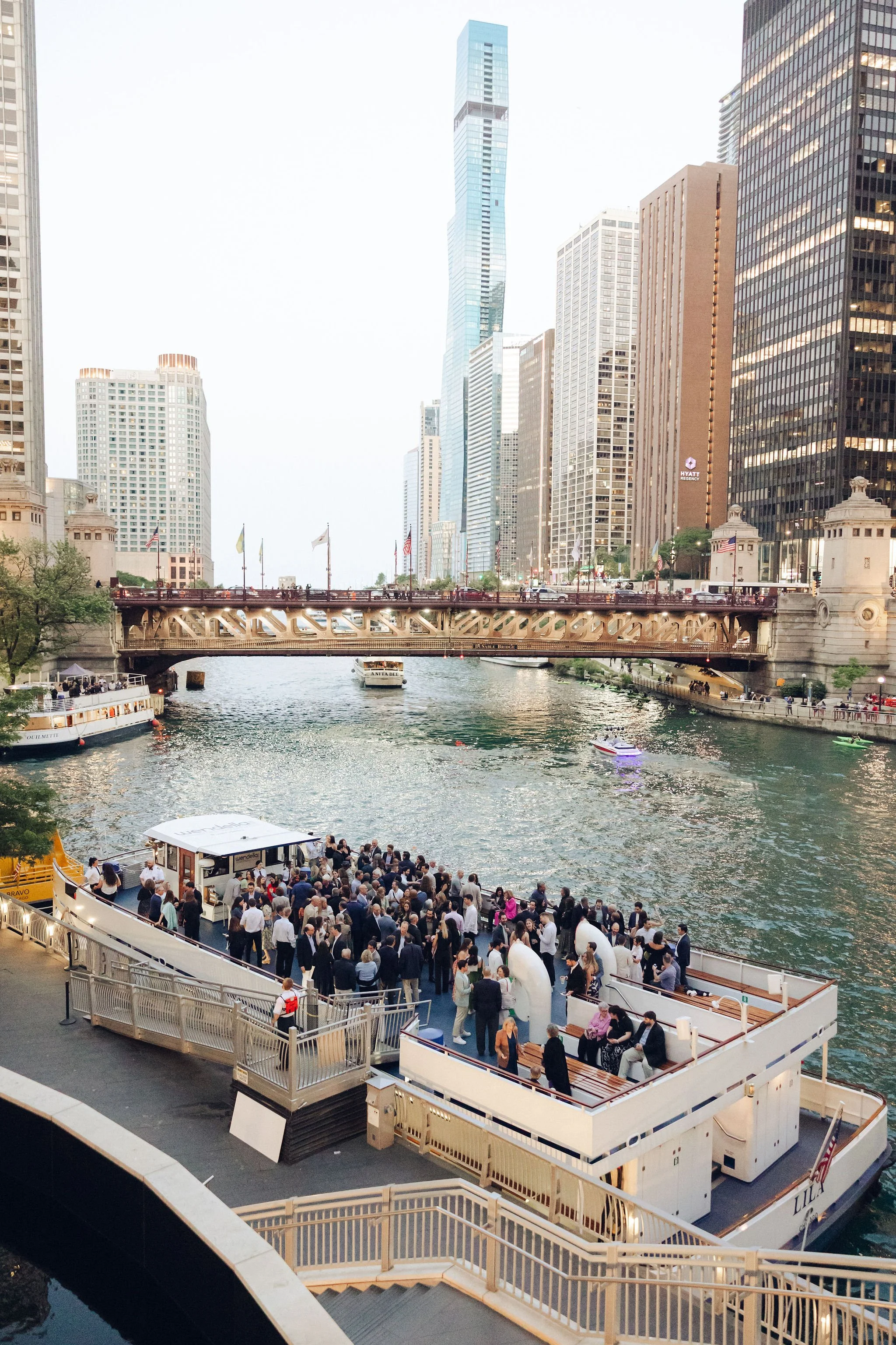 Crowd gathering on a boat docked along a city riverfront, with tall skyscrapers and a bridge in the background.