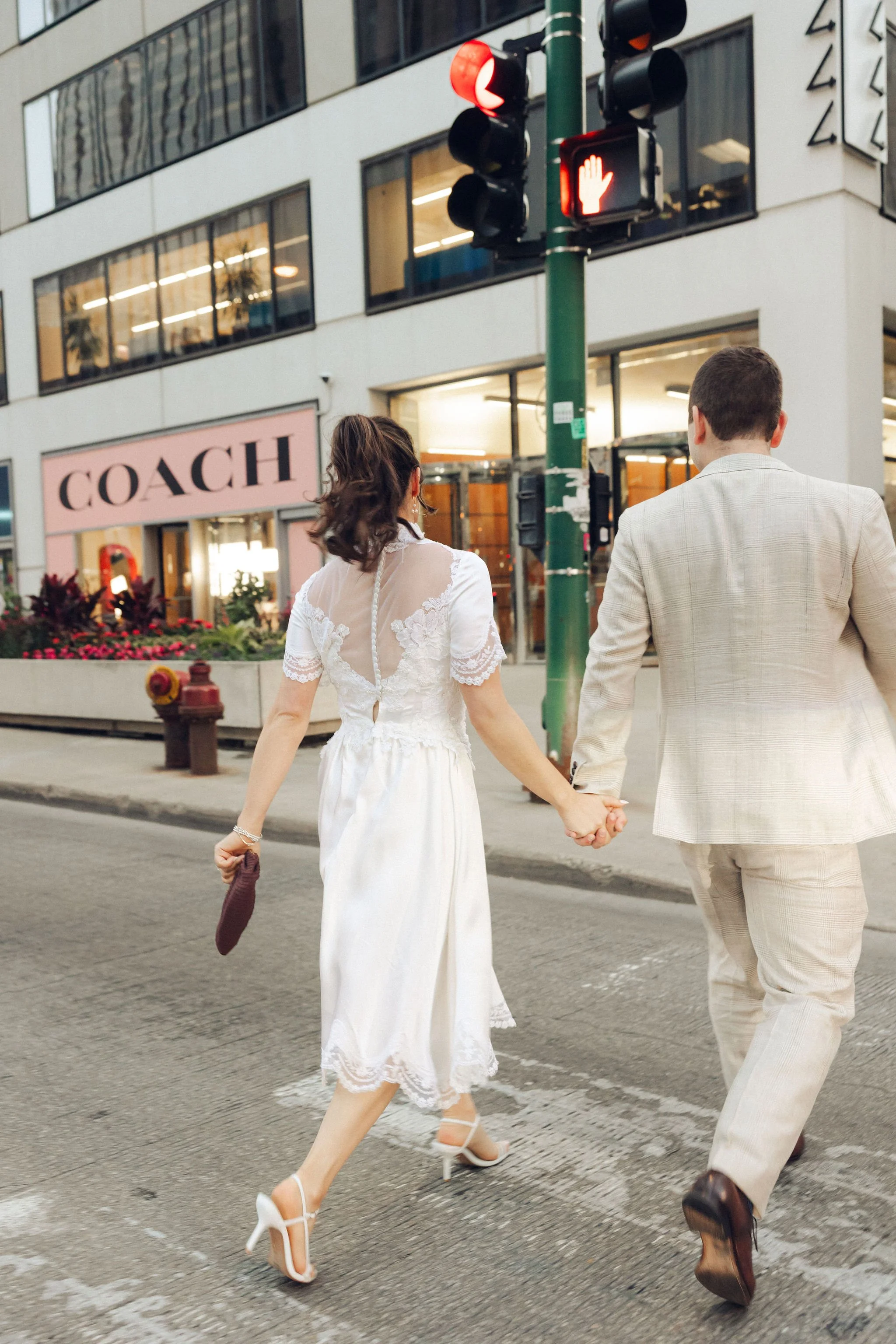 A couple holding hands while crossing a city street at night, with a fashion boutique named Coach visible in the background.