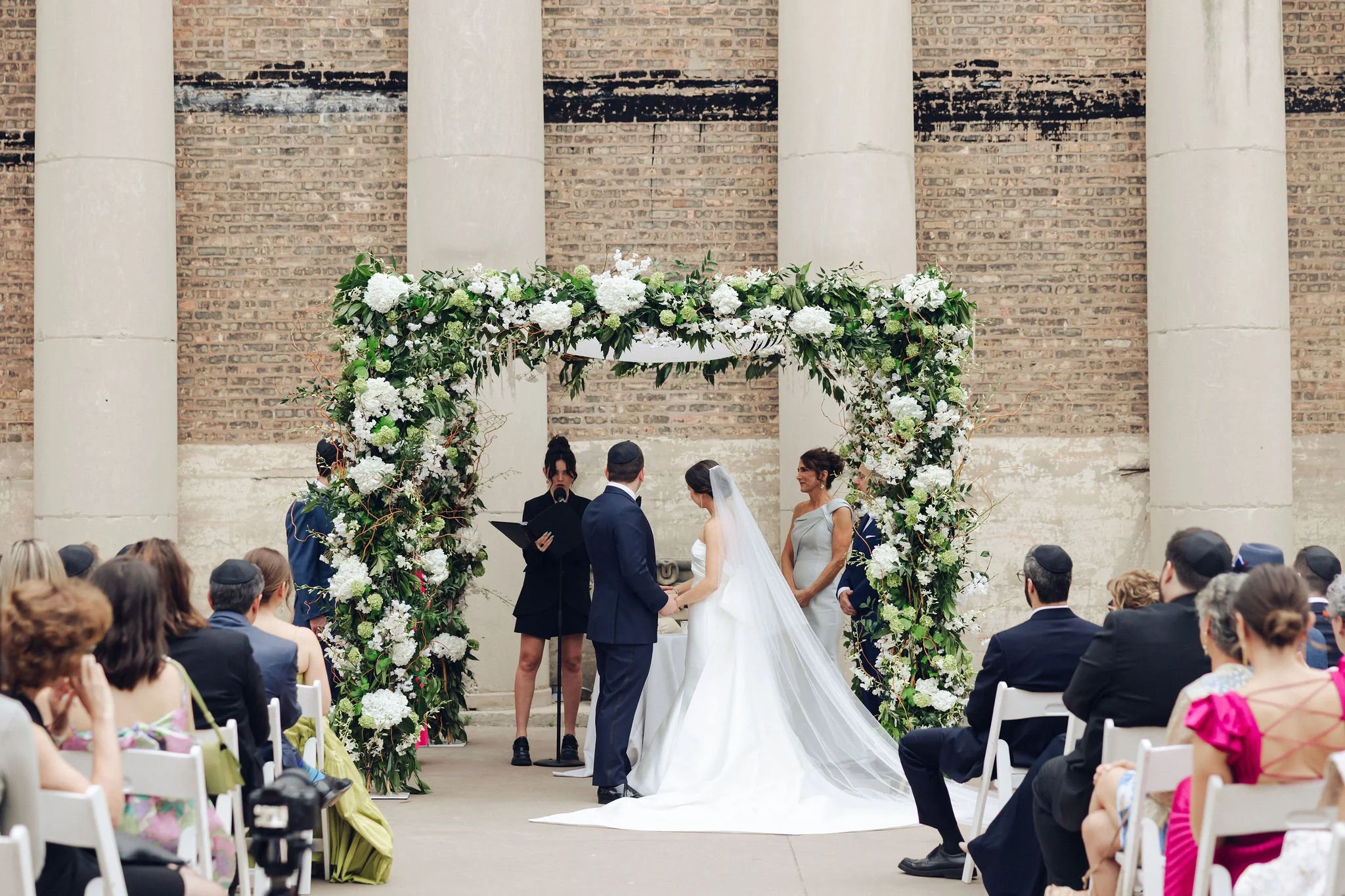 A wedding ceremony taking place outdoors under a floral arch with white flowers and greenery. The bride and groom are holding hands, facing each other, with the officiant and a woman, possibly a family member or bridesmaid, nearby. Guests are seated