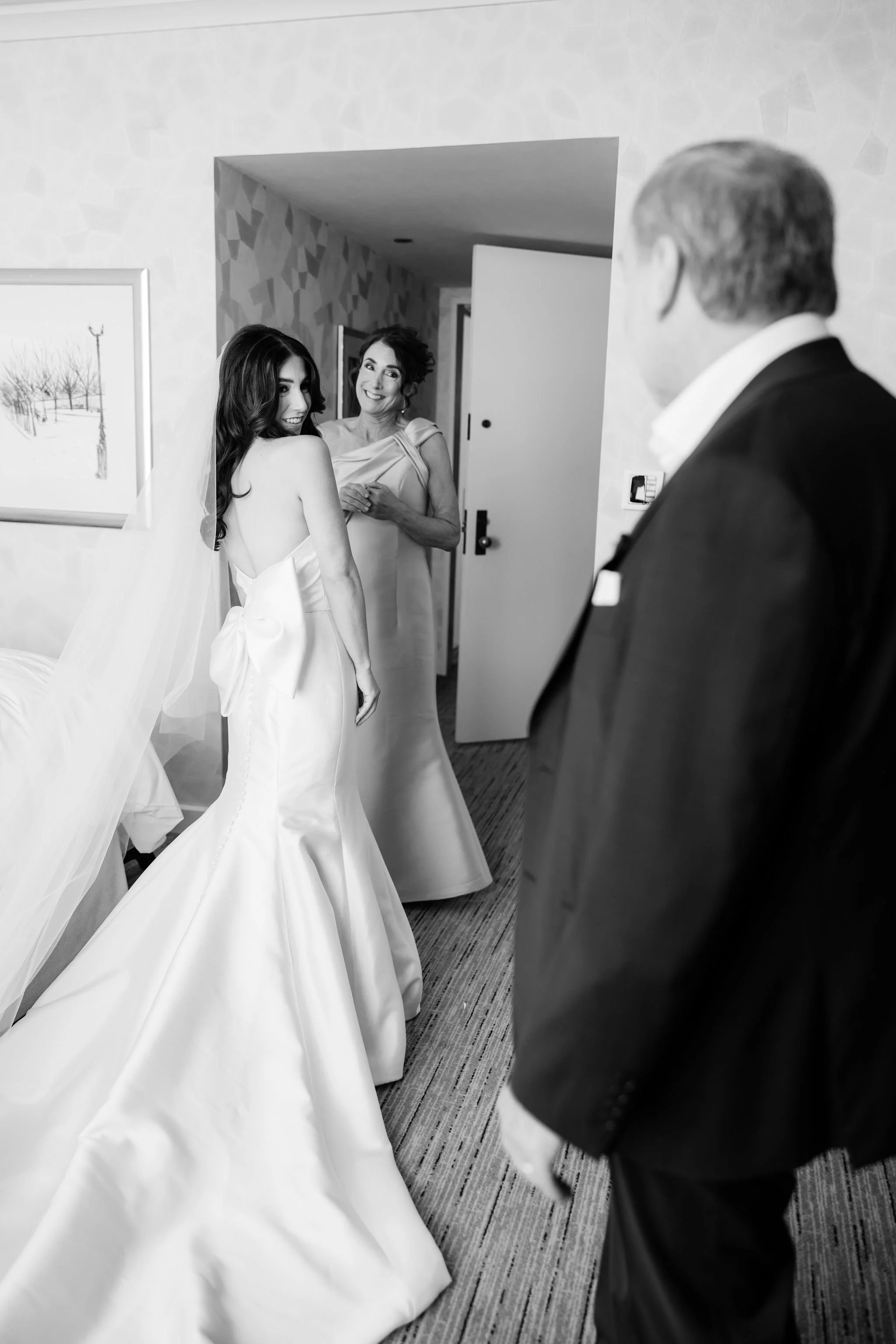 A bride in a wedding dress looking over her shoulder with a smile, a standing woman in a dress smiling, and a man in a tuxedo observing in a hotel room.