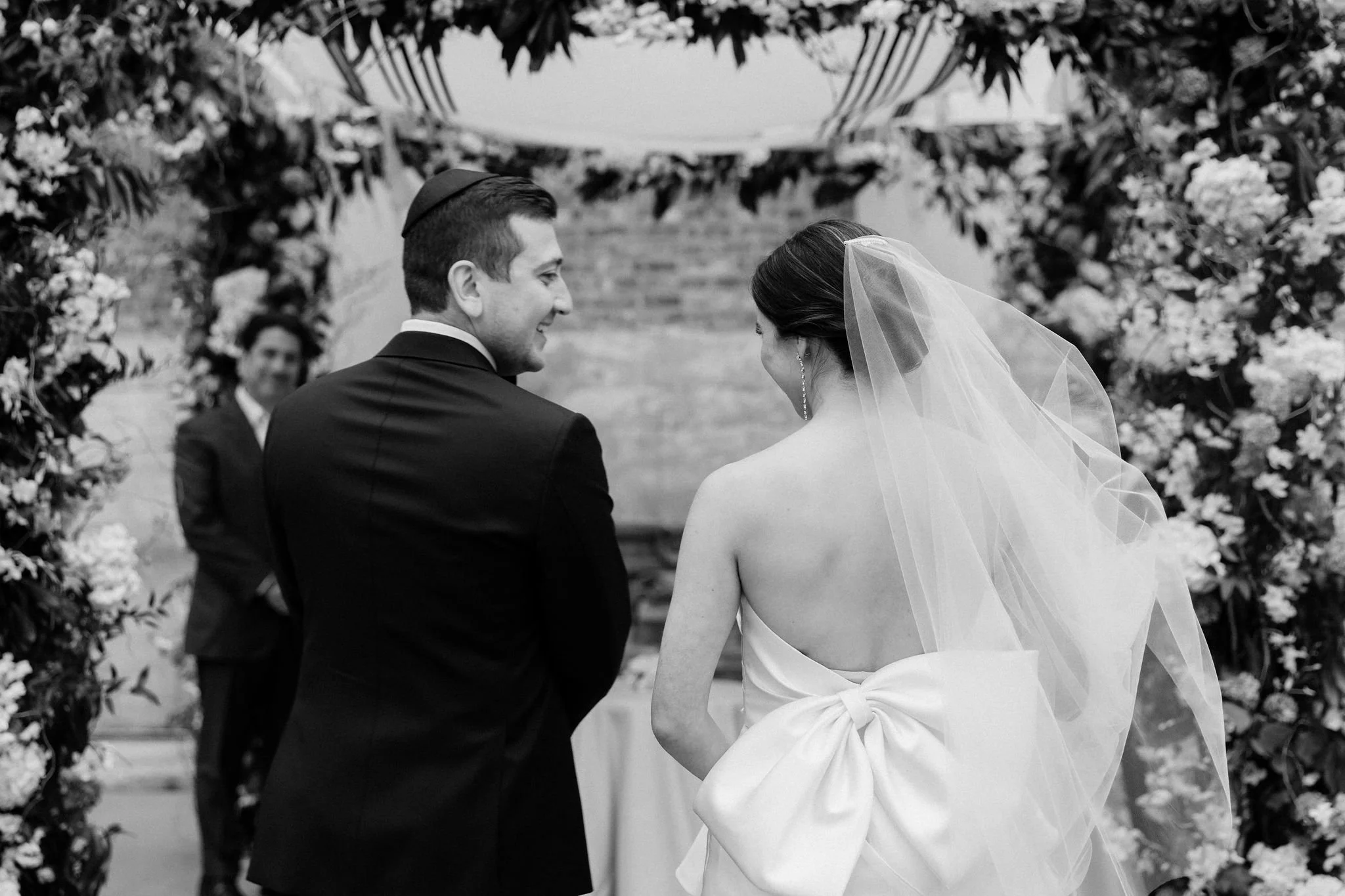A black-and-white photo of a bride and groom during their wedding ceremony, standing under a floral arch, with a person in the background.