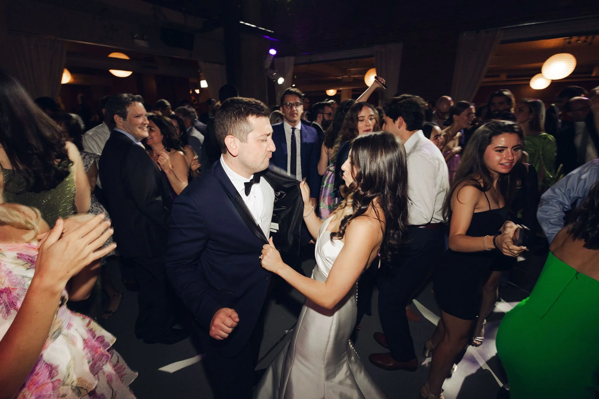People dancing at a party or wedding reception, with a couple in wedding attire in the center.