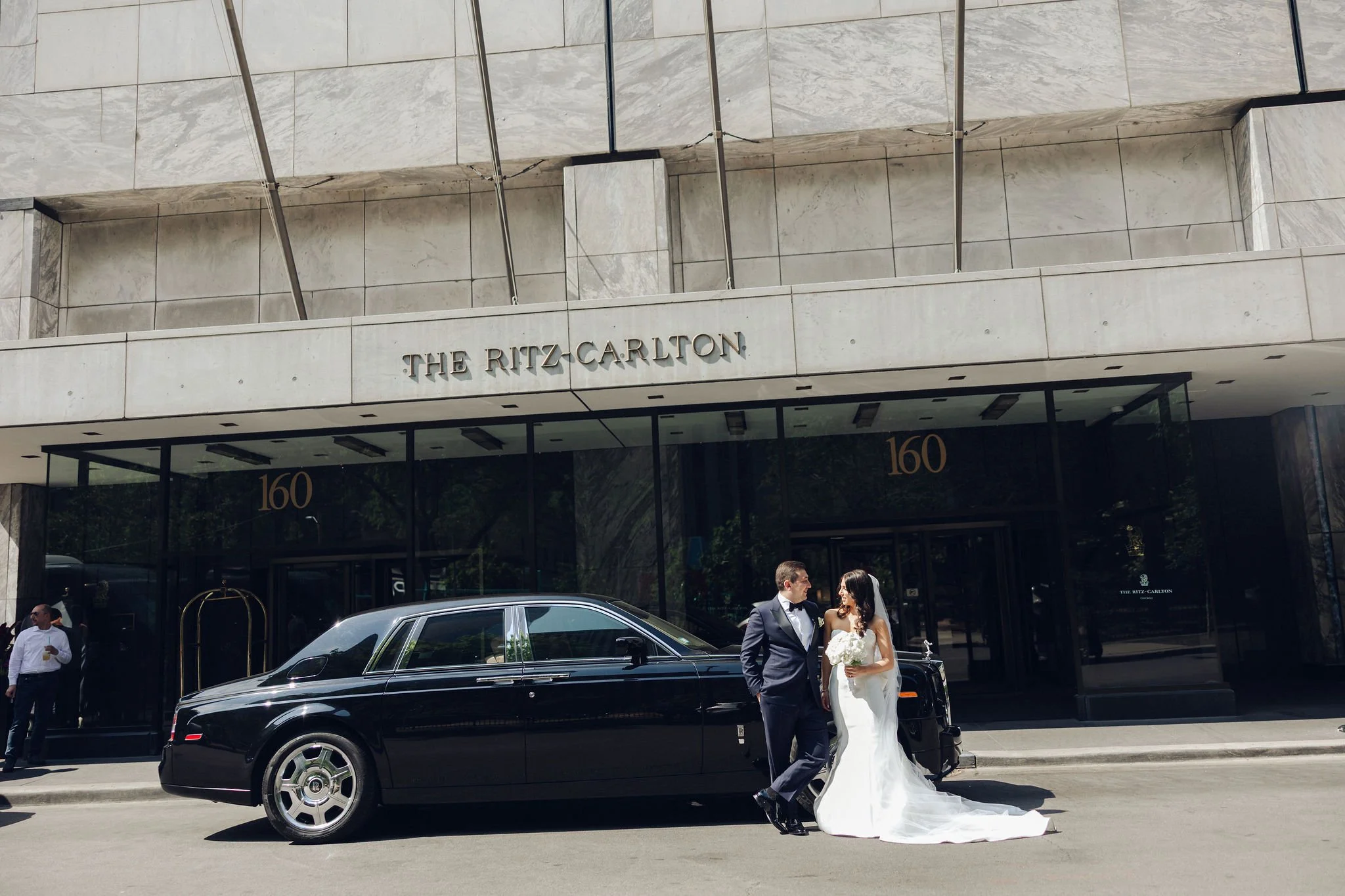 Bride and groom standing in front of The Ritz-Carlton hotel with a black luxury car.