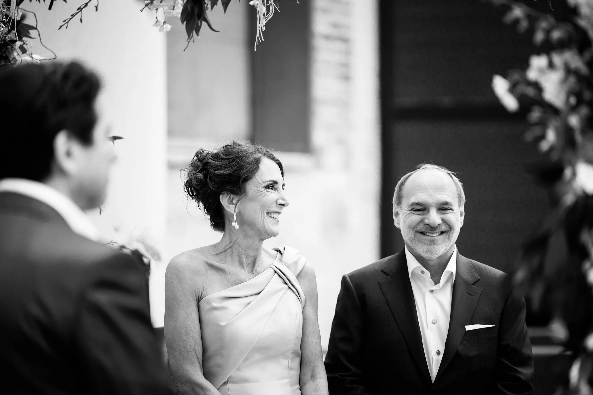 A black-and-white photo of three people at a wedding or formal event, with a woman in an elegant dress smiling and talking to a man in a suit, with a blurred person on the left and floral decorations framing the scene.