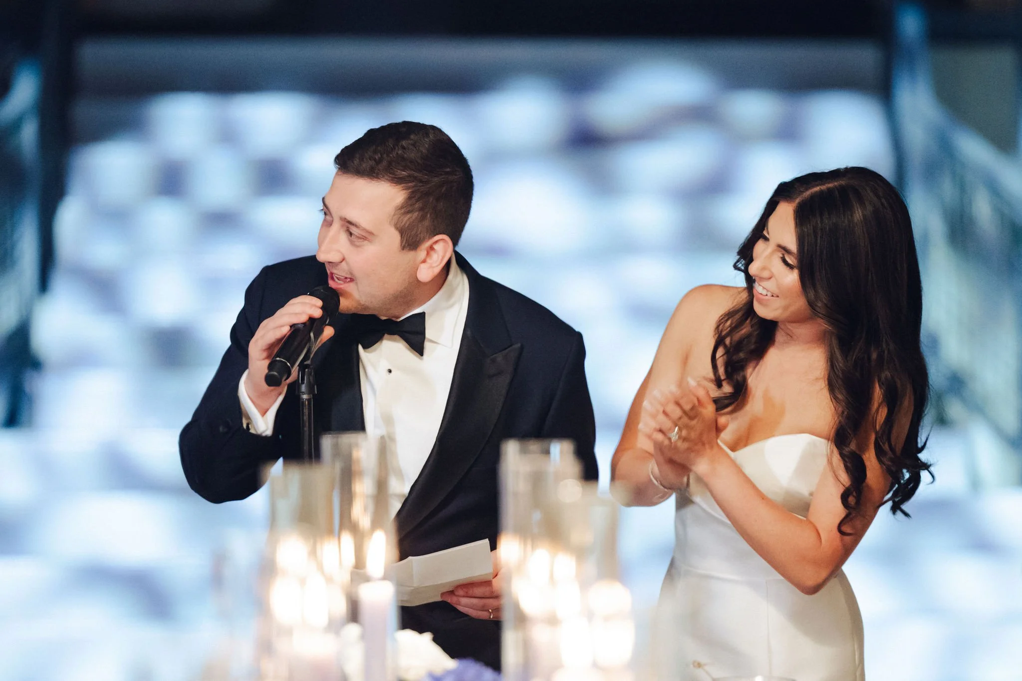 A groom in a black tuxedo with a bow tie is speaking into a microphone, standing next to a smiling bride in a strapless white wedding dress, at their wedding reception with lit candles on the table in front of them.