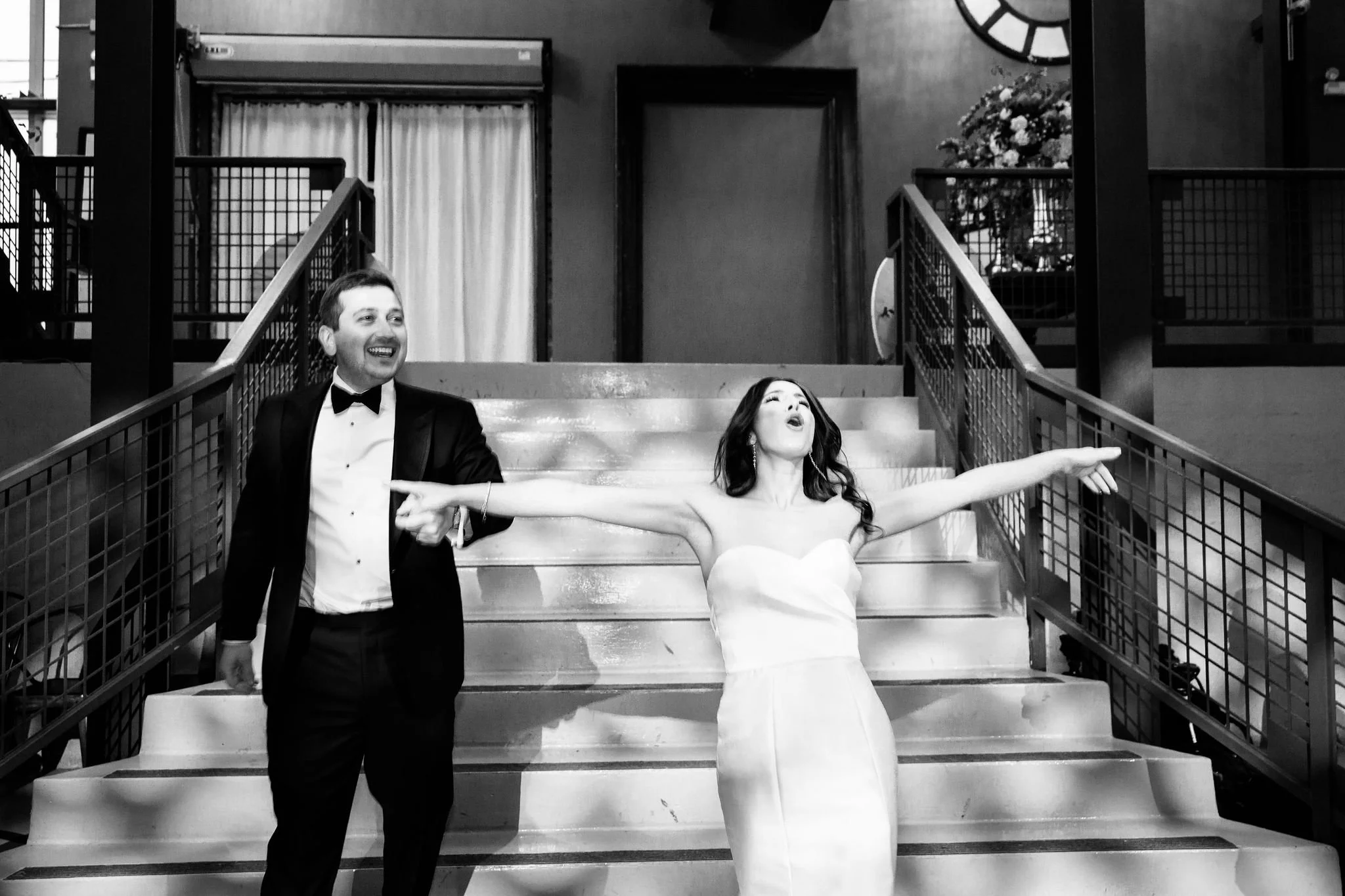 A woman in a wedding dress and a man in a tuxedo walk down stairs, with her arms outstretched and her head tilted back, smiling and appearing joyful in a black and white photo.