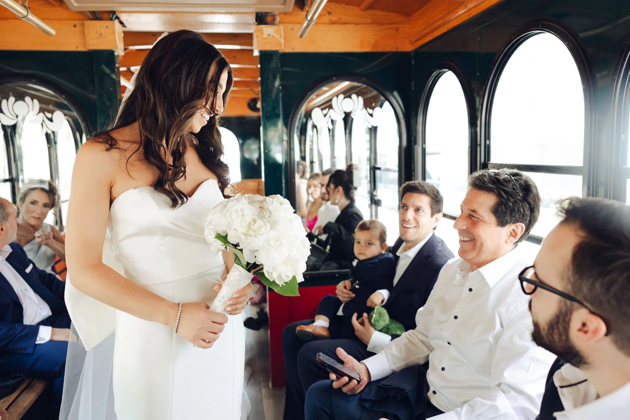 A bride in a white wedding dress holding a bouquet of white flowers inside a train carriage with smiling wedding guests seated along the sides