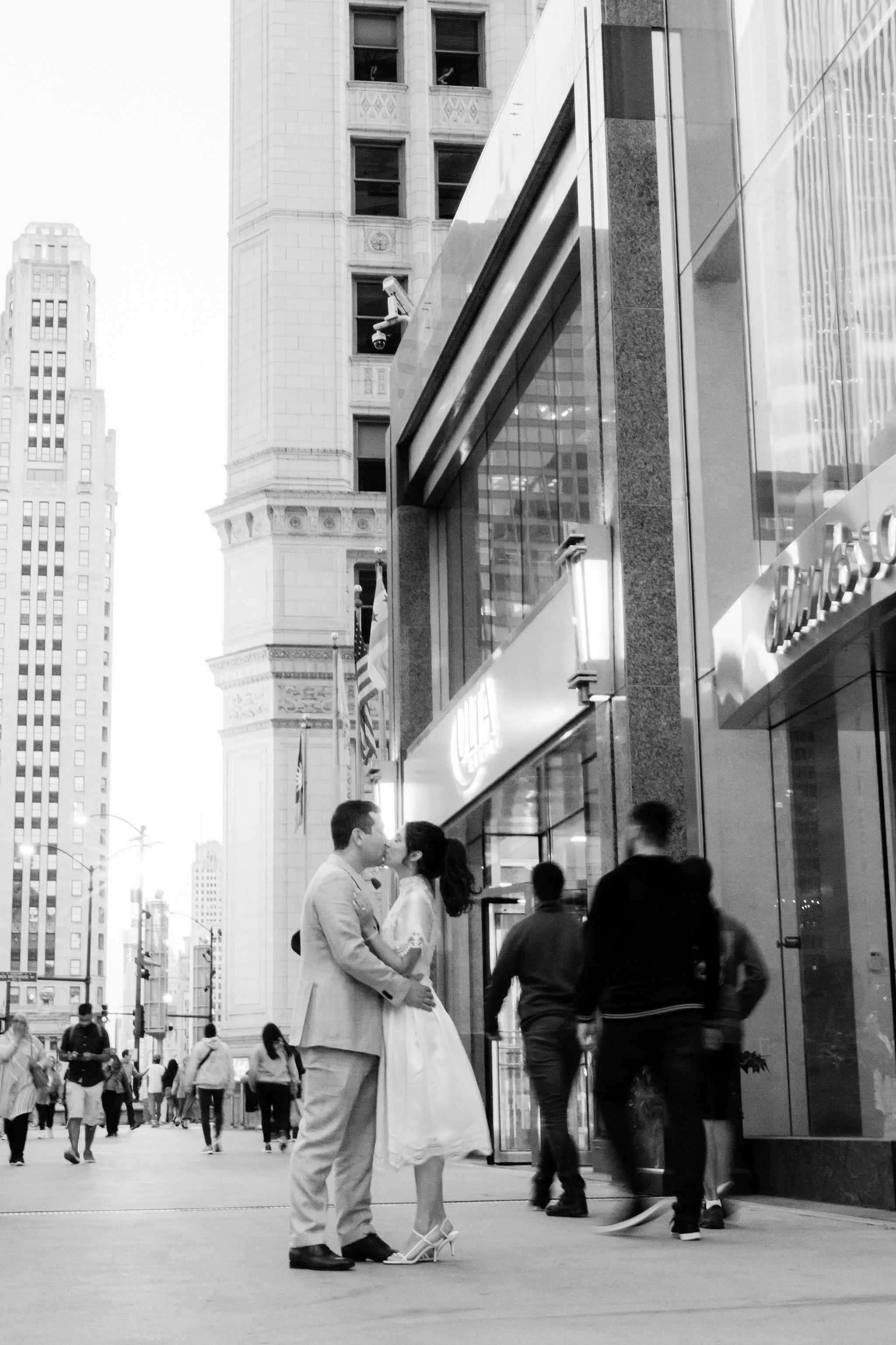 A black and white photo of a city street with a couple kissing in the foreground. The woman is wearing a dress and heels, and the man is in a suit. There are pedestrians walking past and tall buildings in the background.