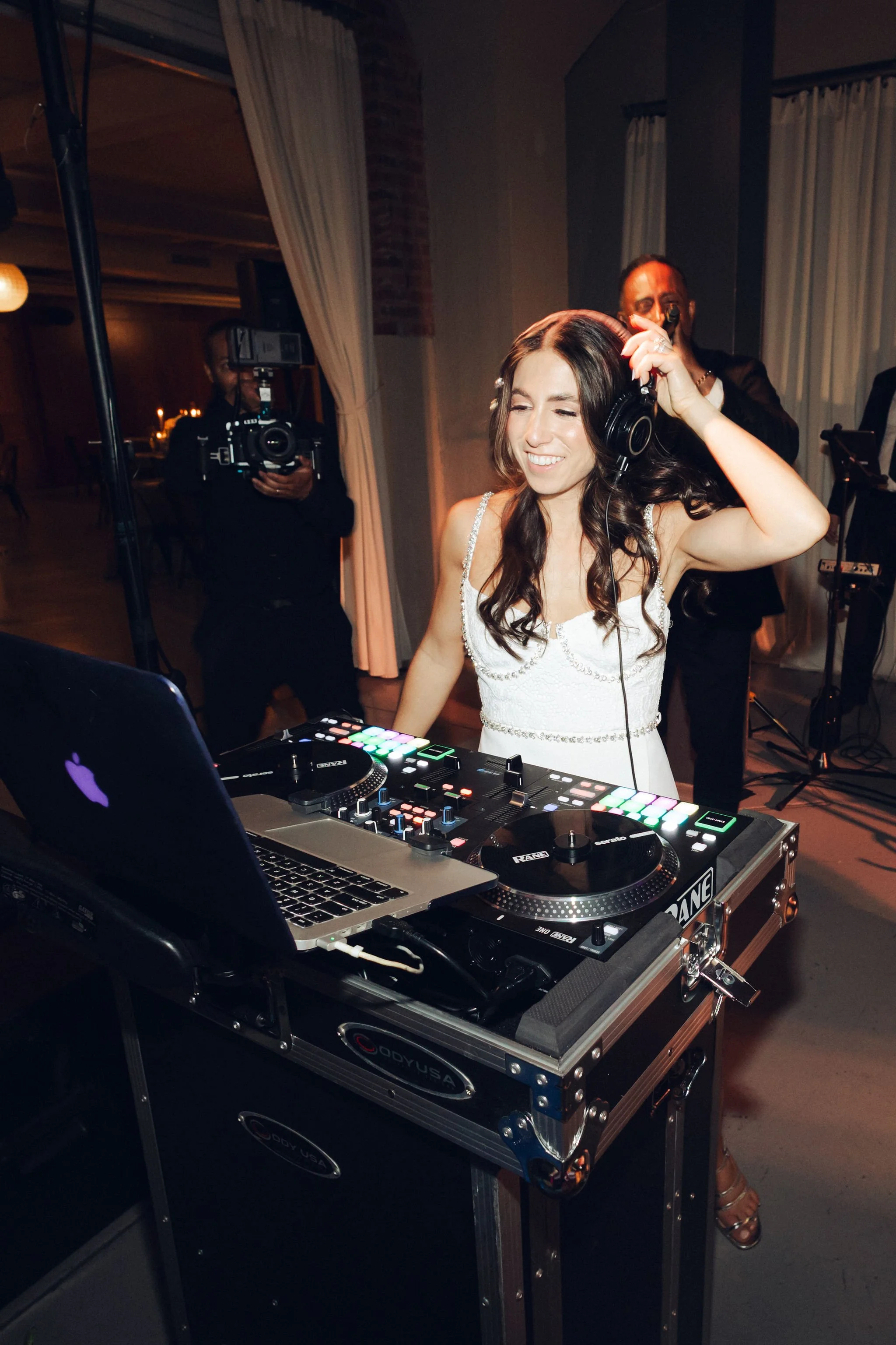 A woman in a white dress is DJing at an event, smiling, with headphones on, in front of DJ equipment and a laptop, while a man videotapes her in the background.