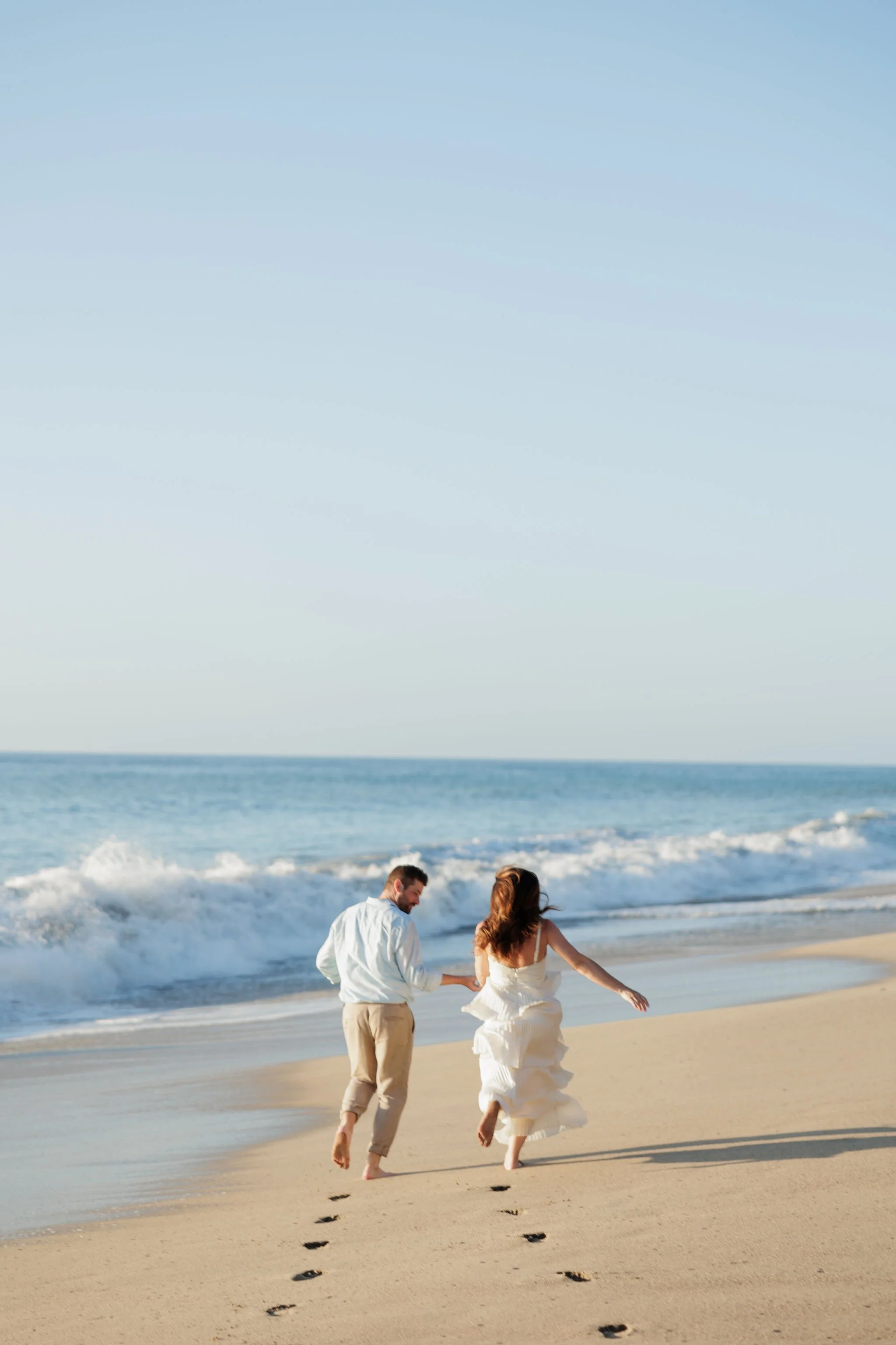 A couple in wedding attire running on the beach, leaving footprints in the sand with the ocean and sky in the background.