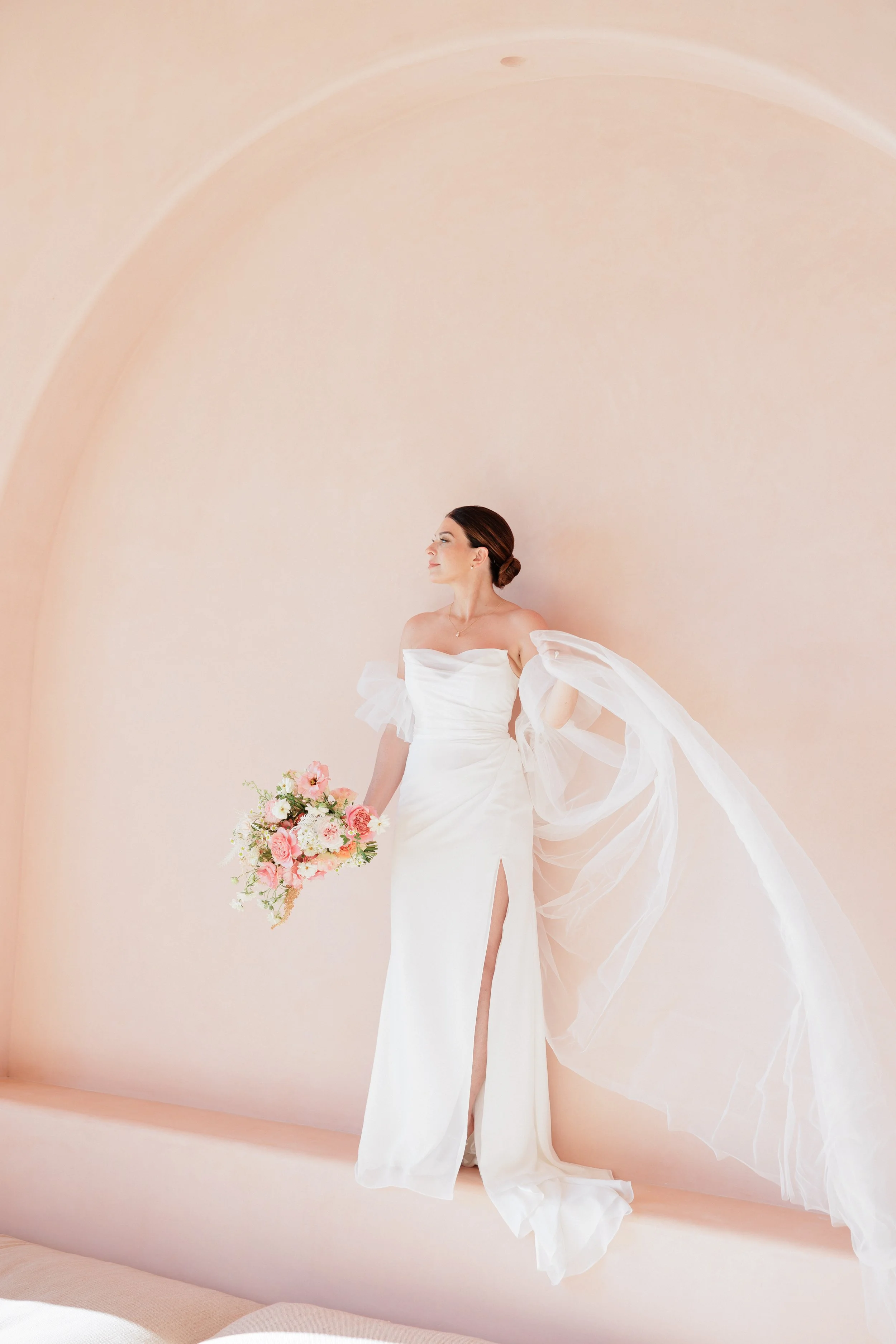 A woman in a white wedding gown holding a bouquet of pink and white flowers, standing against a nude-colored wall.