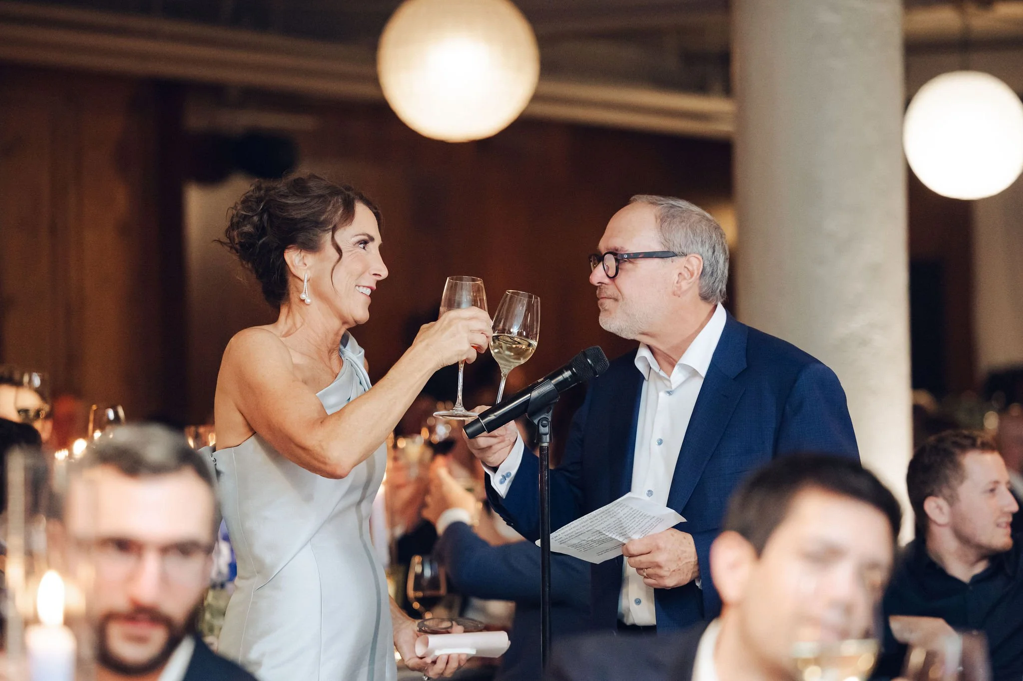 A woman in a sleeveless silver dress and a man in a suit sharing a toast with wine glasses at a formal gathering or celebration.