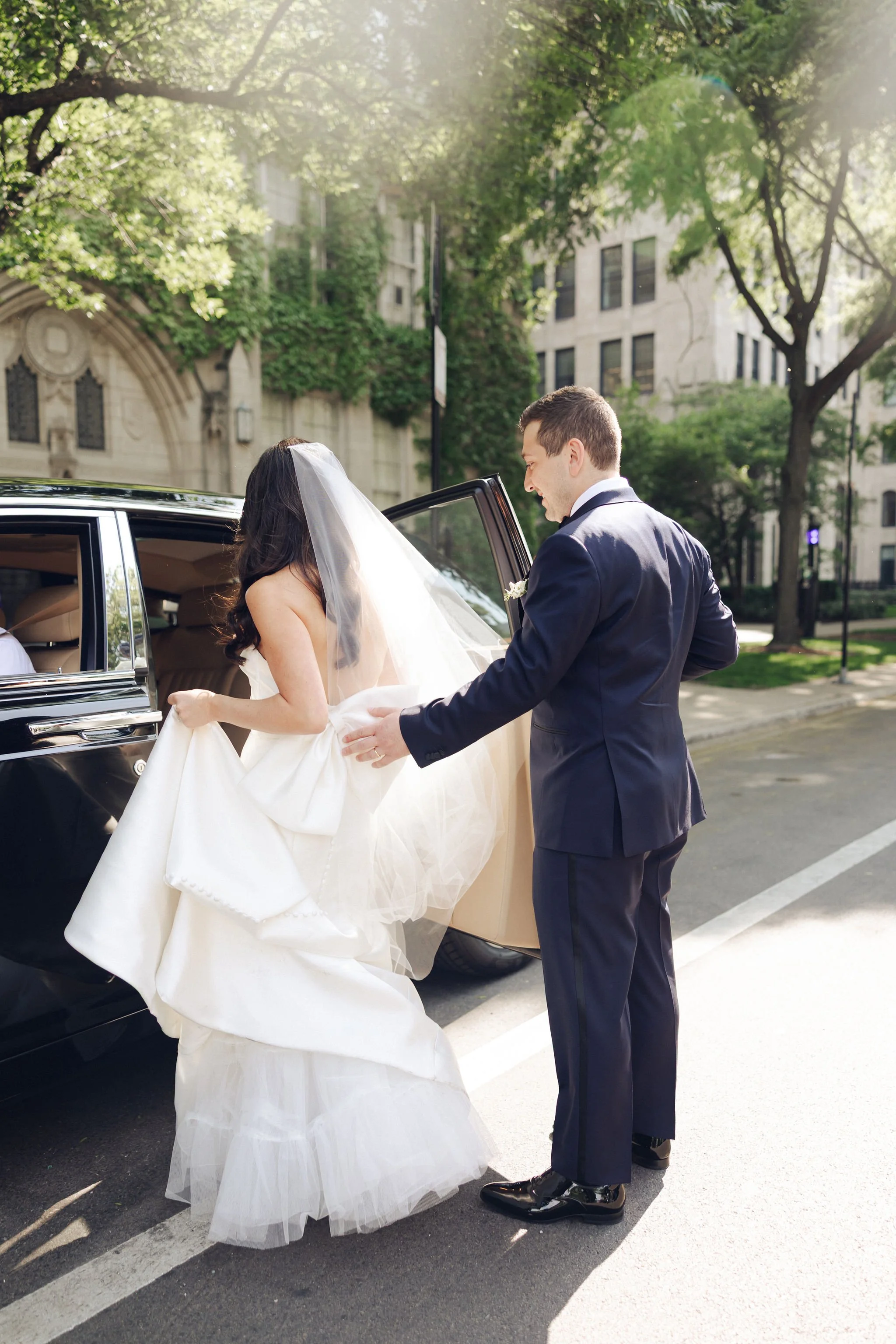 Bride in wedding dress and veil getting into a black limousine with groom in formal suit helping her.