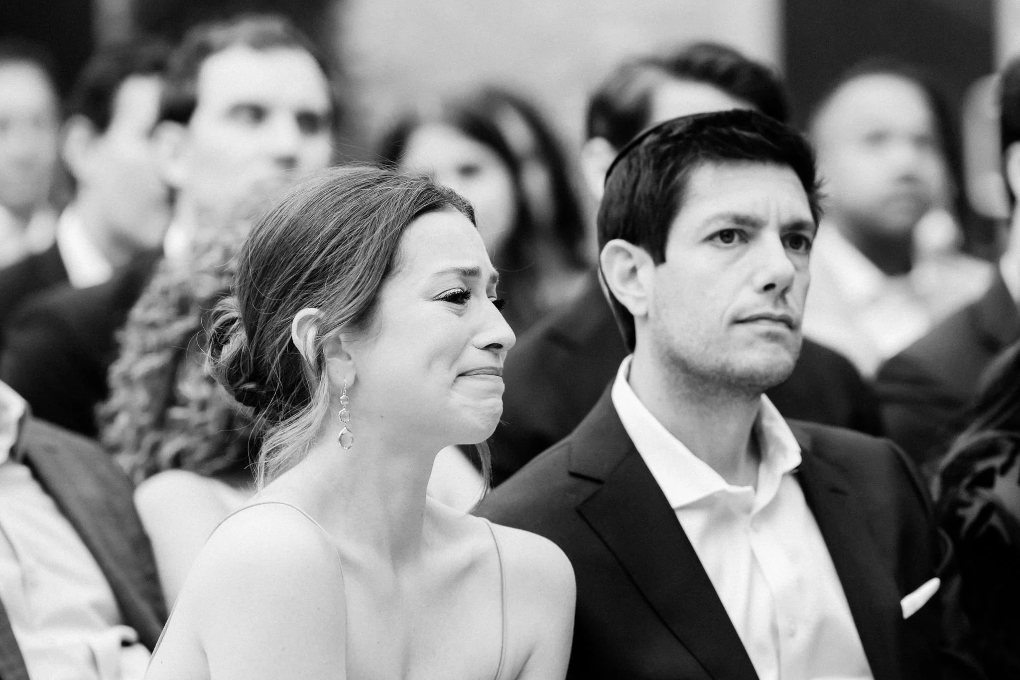 Black and white photo of two people sitting in an audience at a formal event, with other attendees visible in the background.