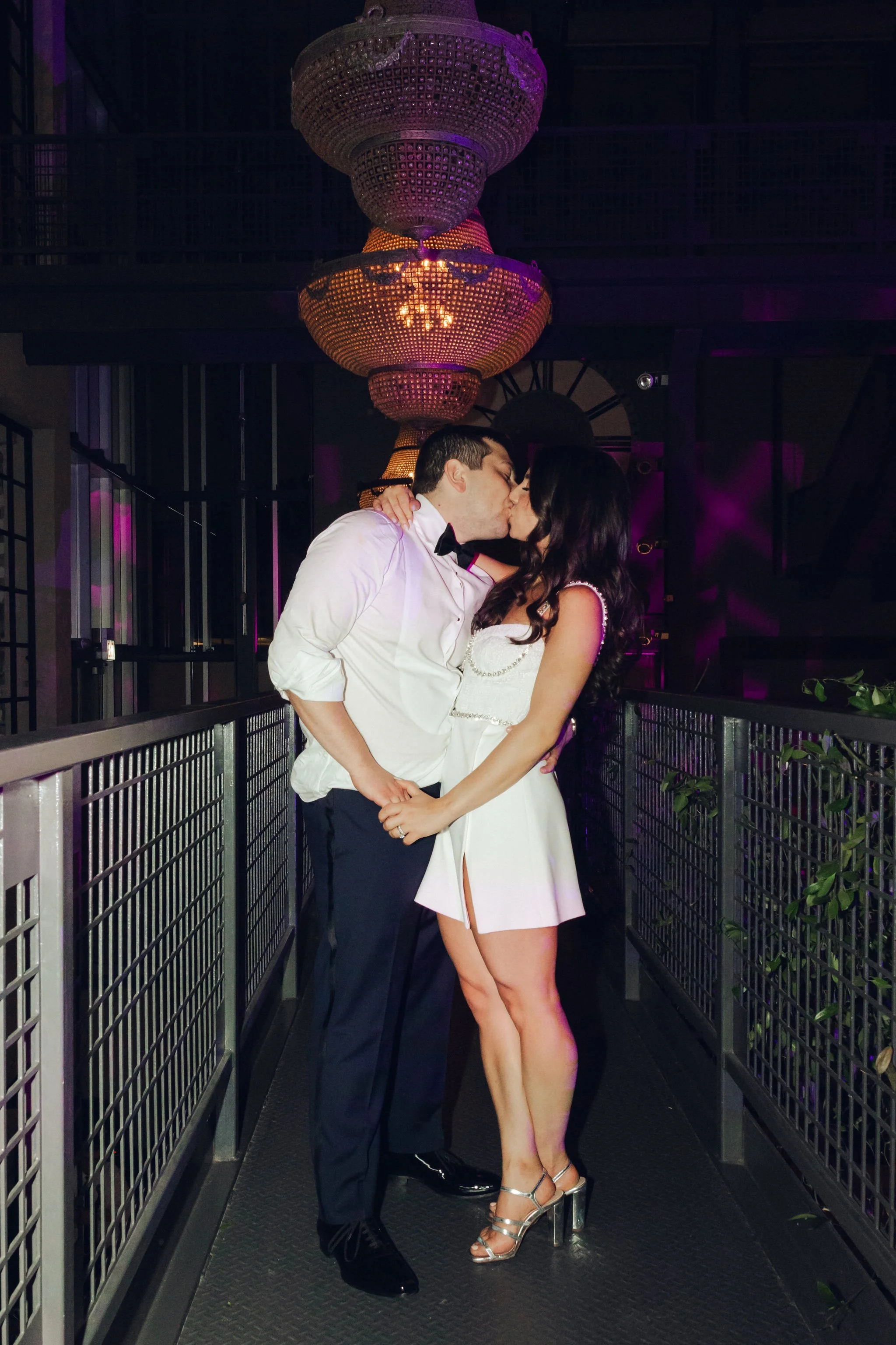 A couple in formal attire sharing a kiss on a balcony with a large chandelier overhead.