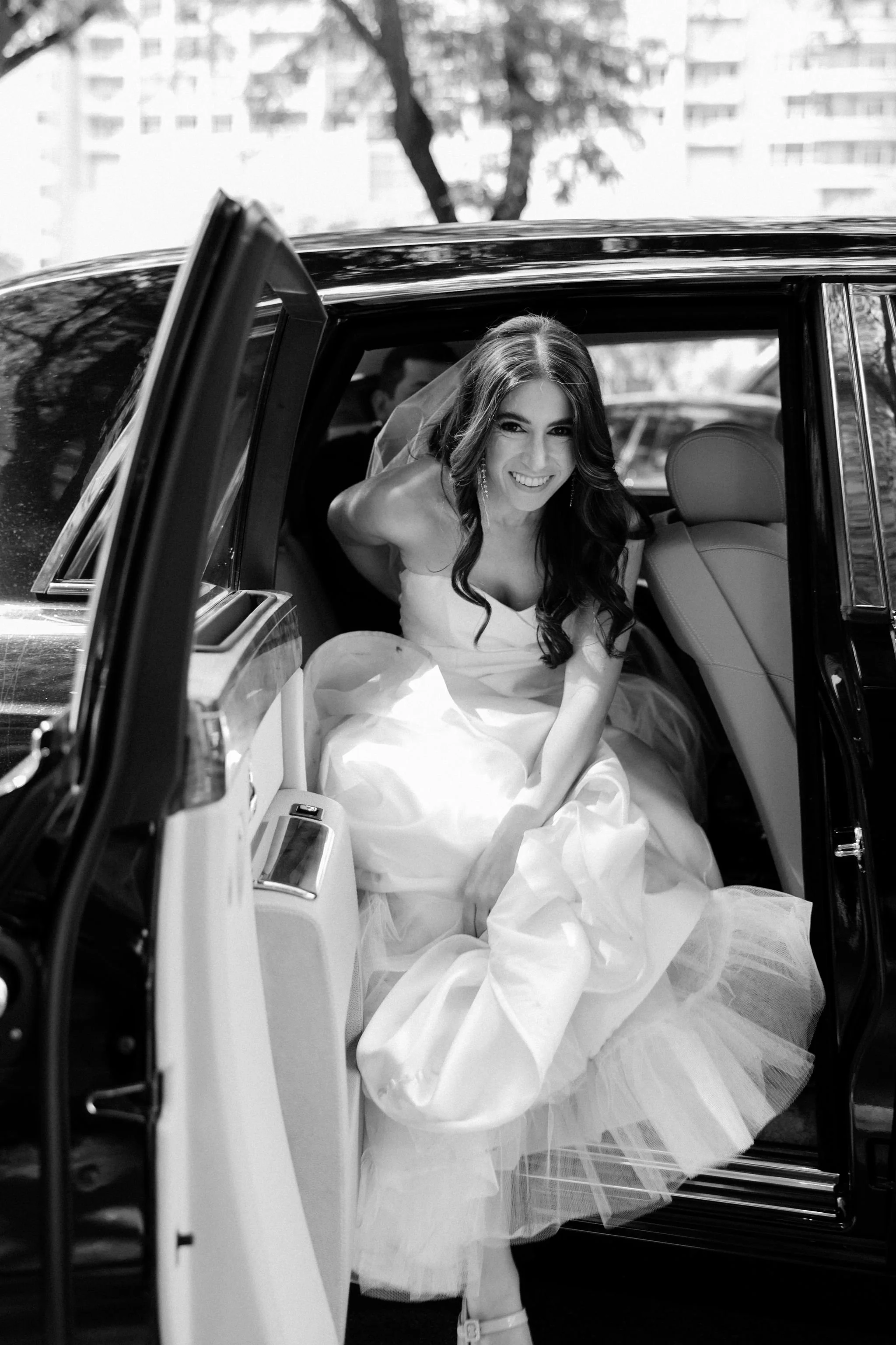 Black-and-white photo of a woman in a wedding dress sitting in the back of a limousine, smiling and looking at the camera.