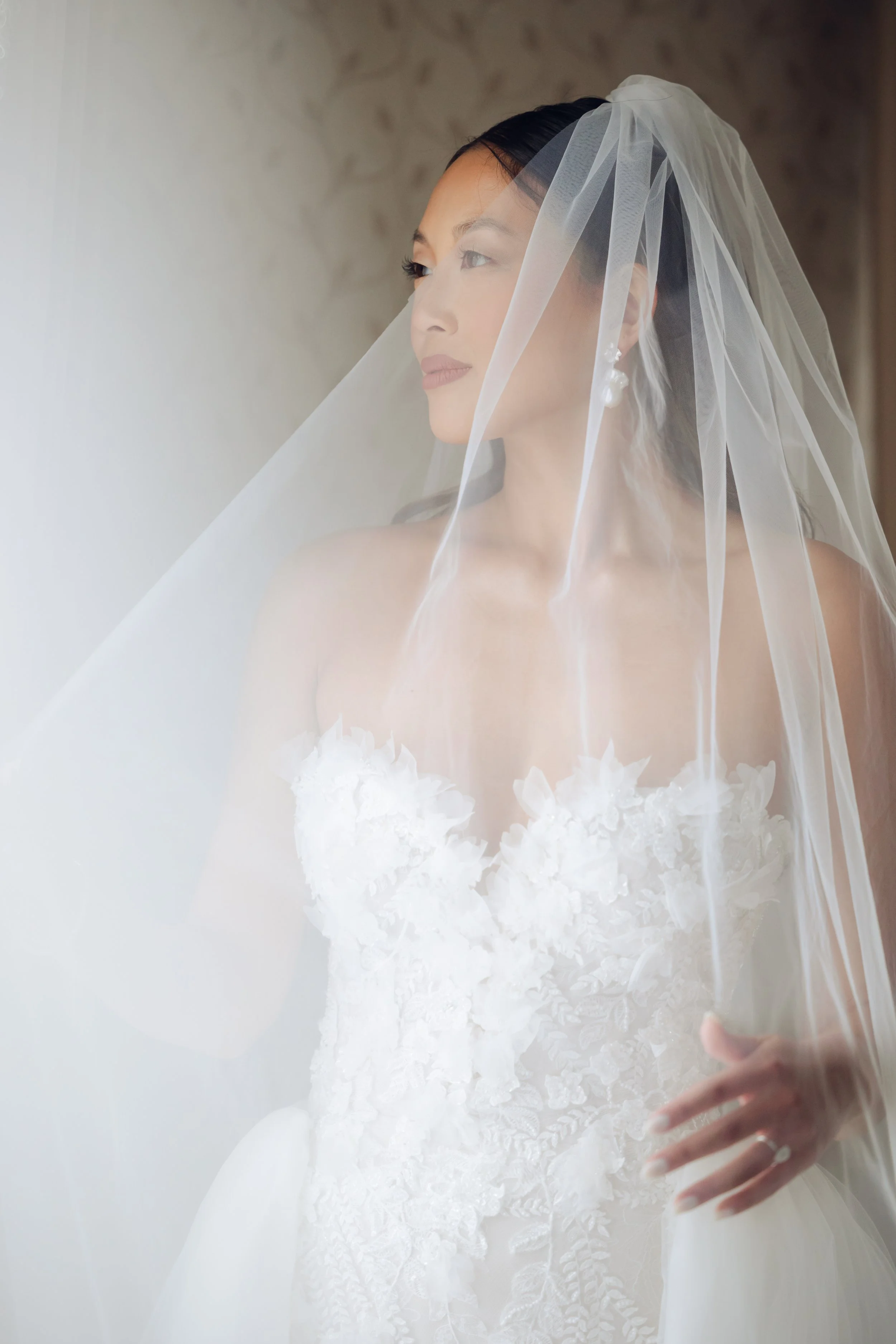 Bride in a white wedding dress with lace details and a veil, looking out a window.