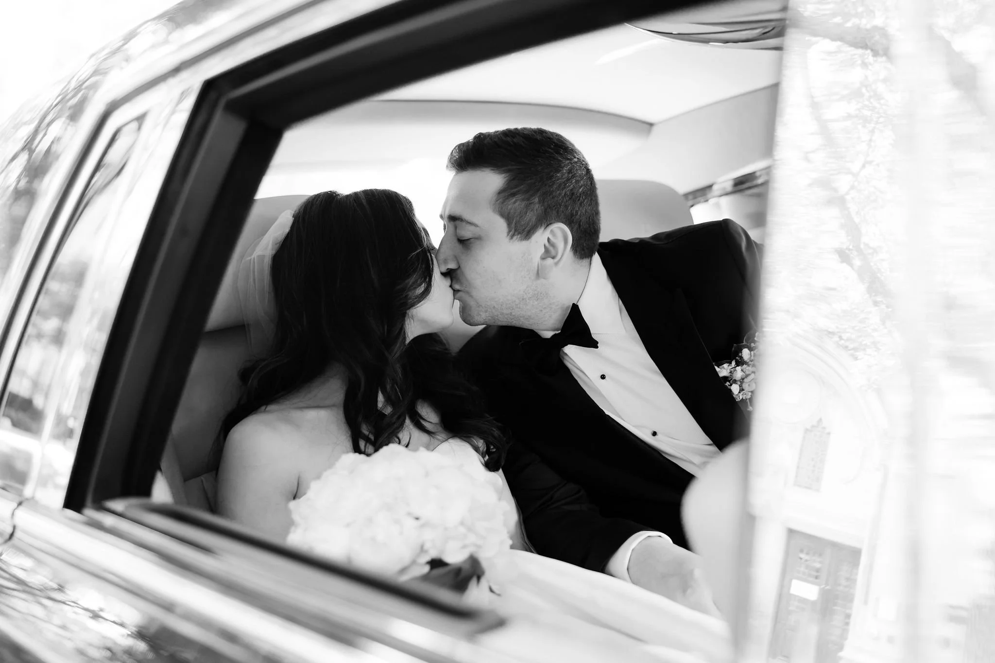 Black and white photo of a newlywed couple sharing a kiss inside a car, with the bride holding a bouquet of flowers.