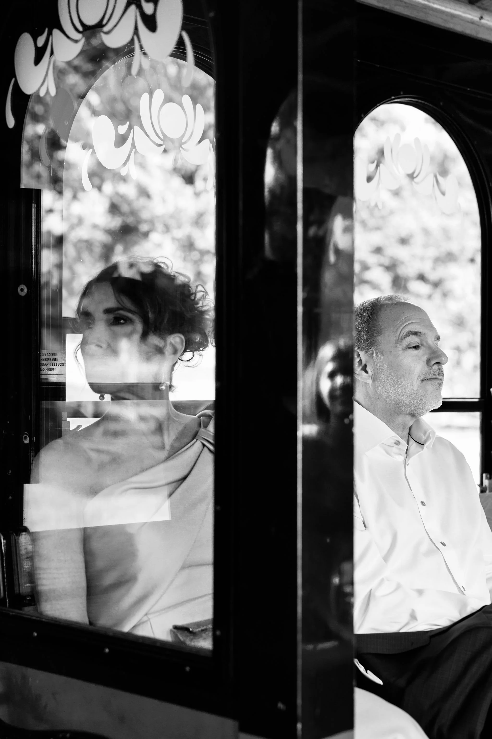 Black and white photo of two people seated inside a vehicle, viewed through a glass window. The woman on the left has short, curly hair and is wearing a light-colored top, looking out to the left. The man on the right has a receding hairline, mustach