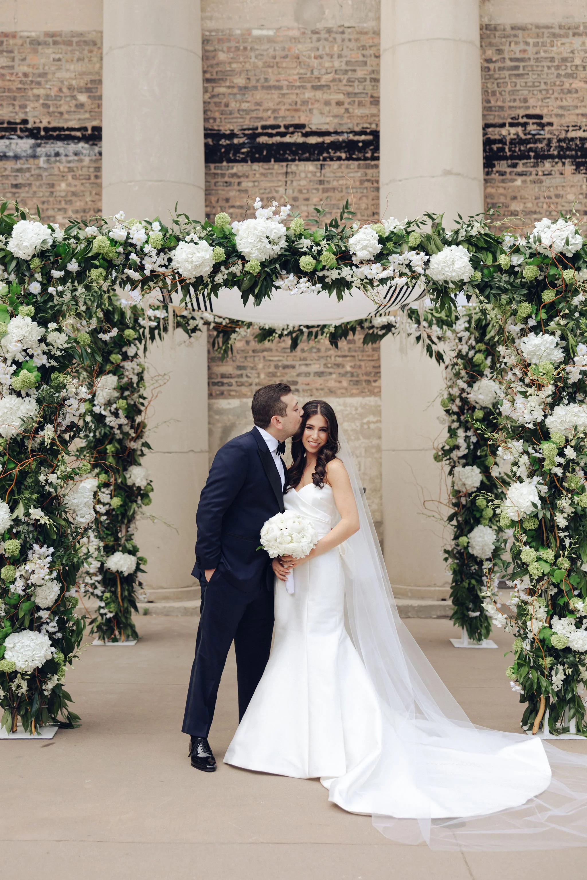 Bride and groom standing under floral arch at outdoor wedding ceremony, bride holding bouquet, groom kissing her cheek, smiling.