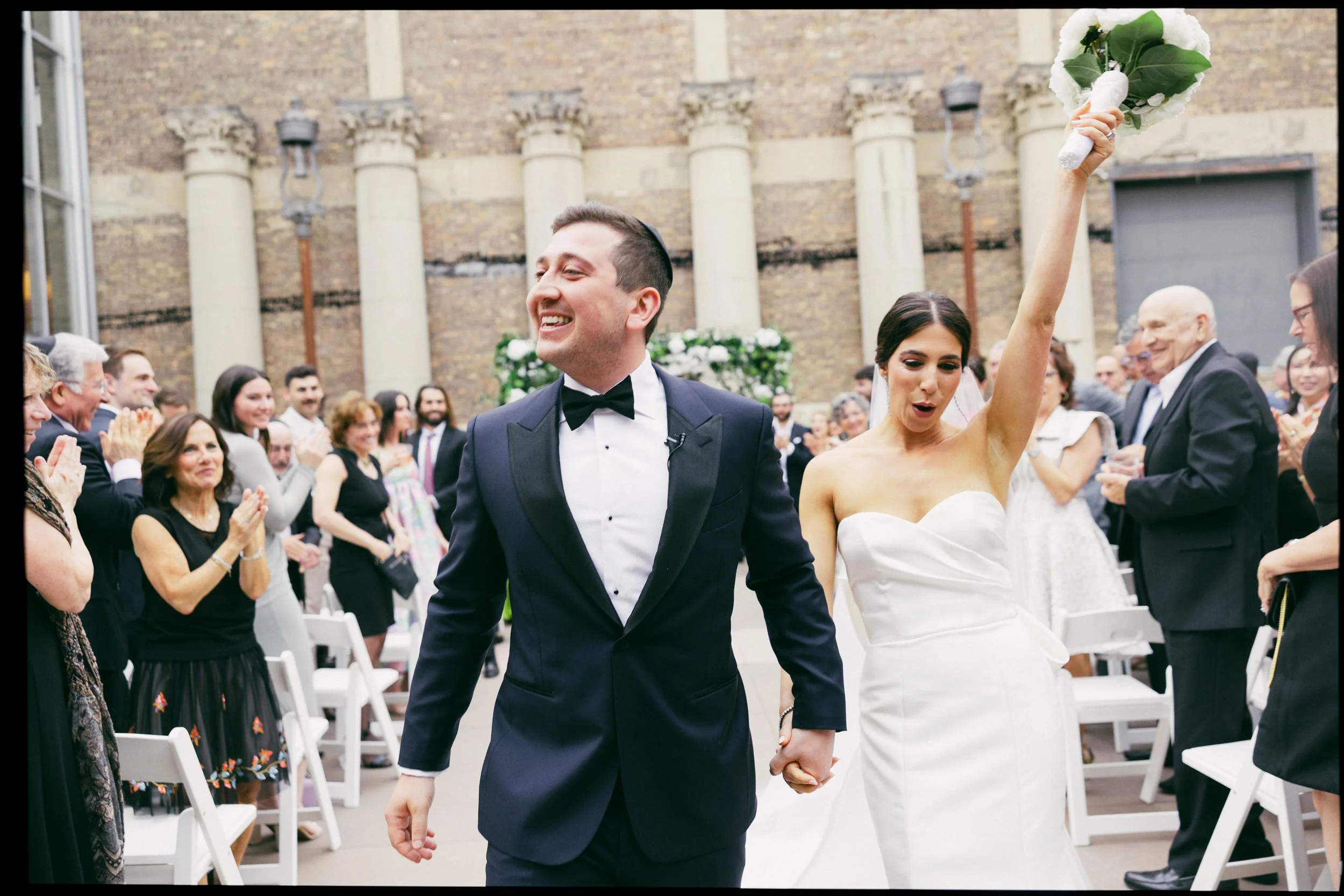 A newly married couple, holding hands, celebrating their wedding before a gathering of friends and family at an outdoor ceremony. The groom is wearing a black tuxedo and the bride is in a white strapless wedding gown, raising her bouquet in the air w