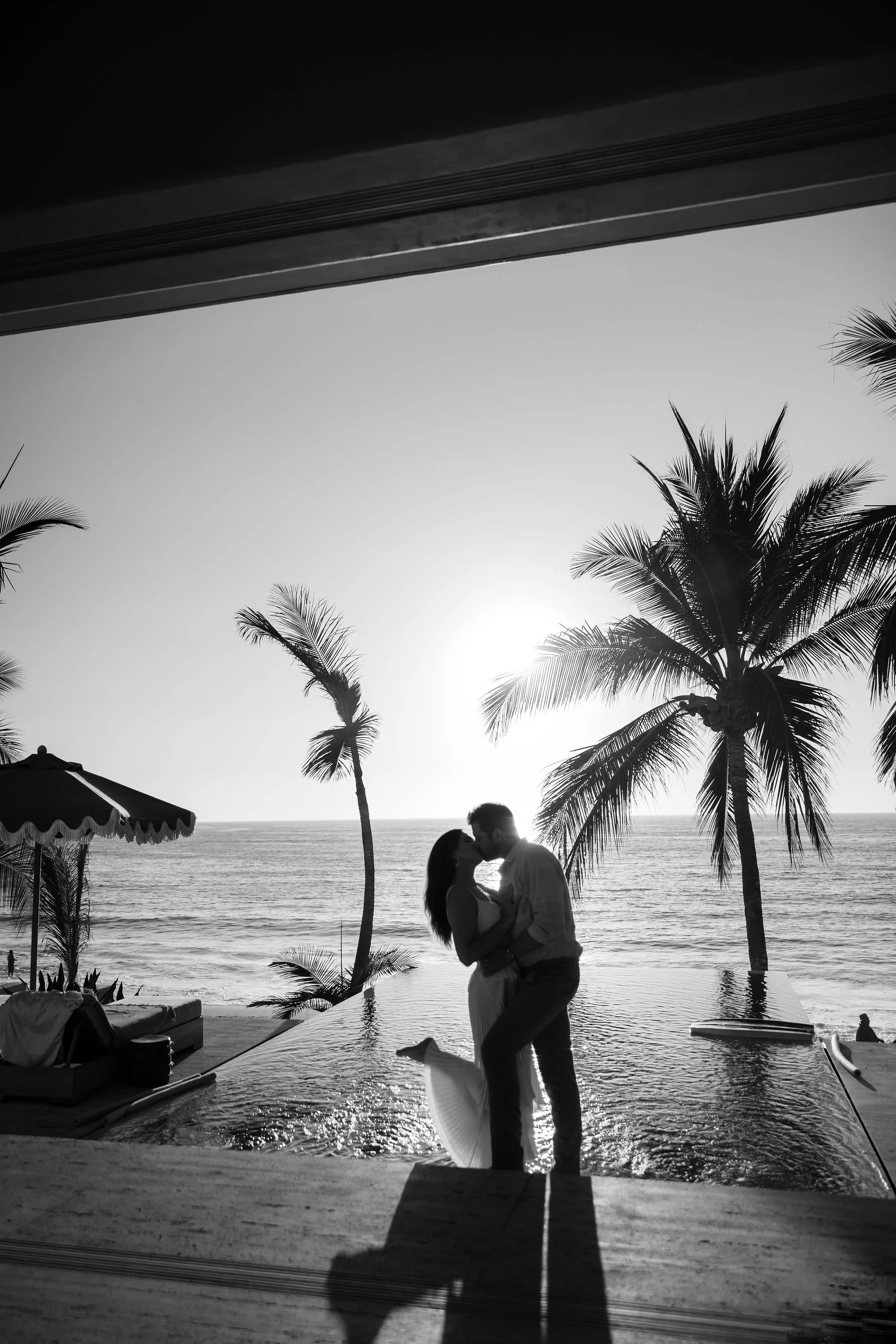 A couple kissing on a beach at sunset with palm trees in the background, seen from a covered outdoor area in black and white.