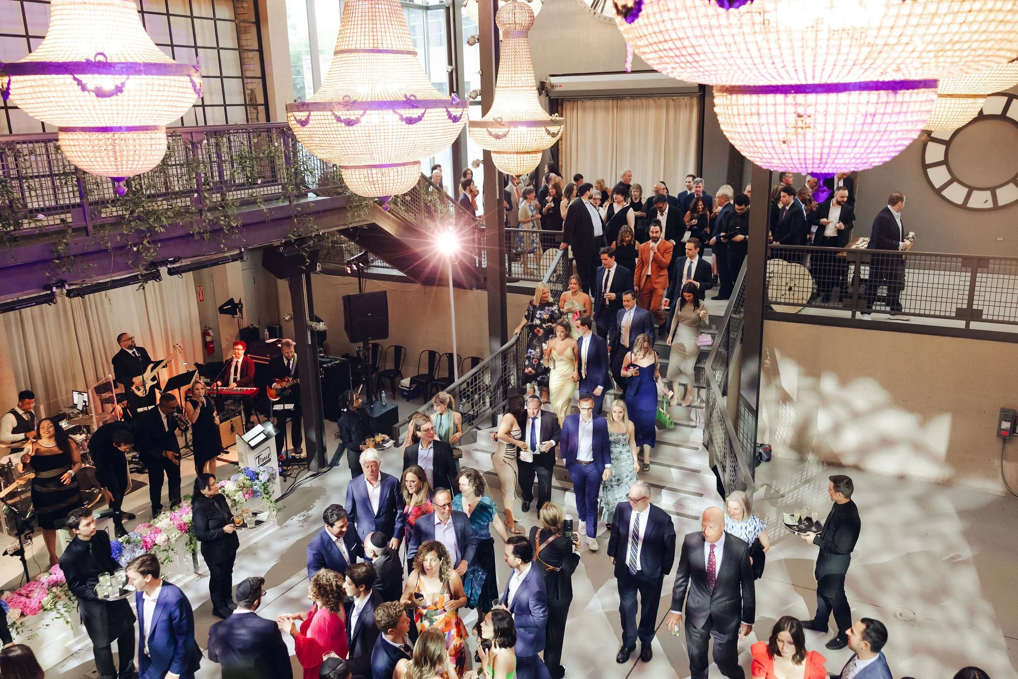 Guests at a formal event dancing and socializing inside a decorated venue with chandeliers, a live band, and floral arrangements.