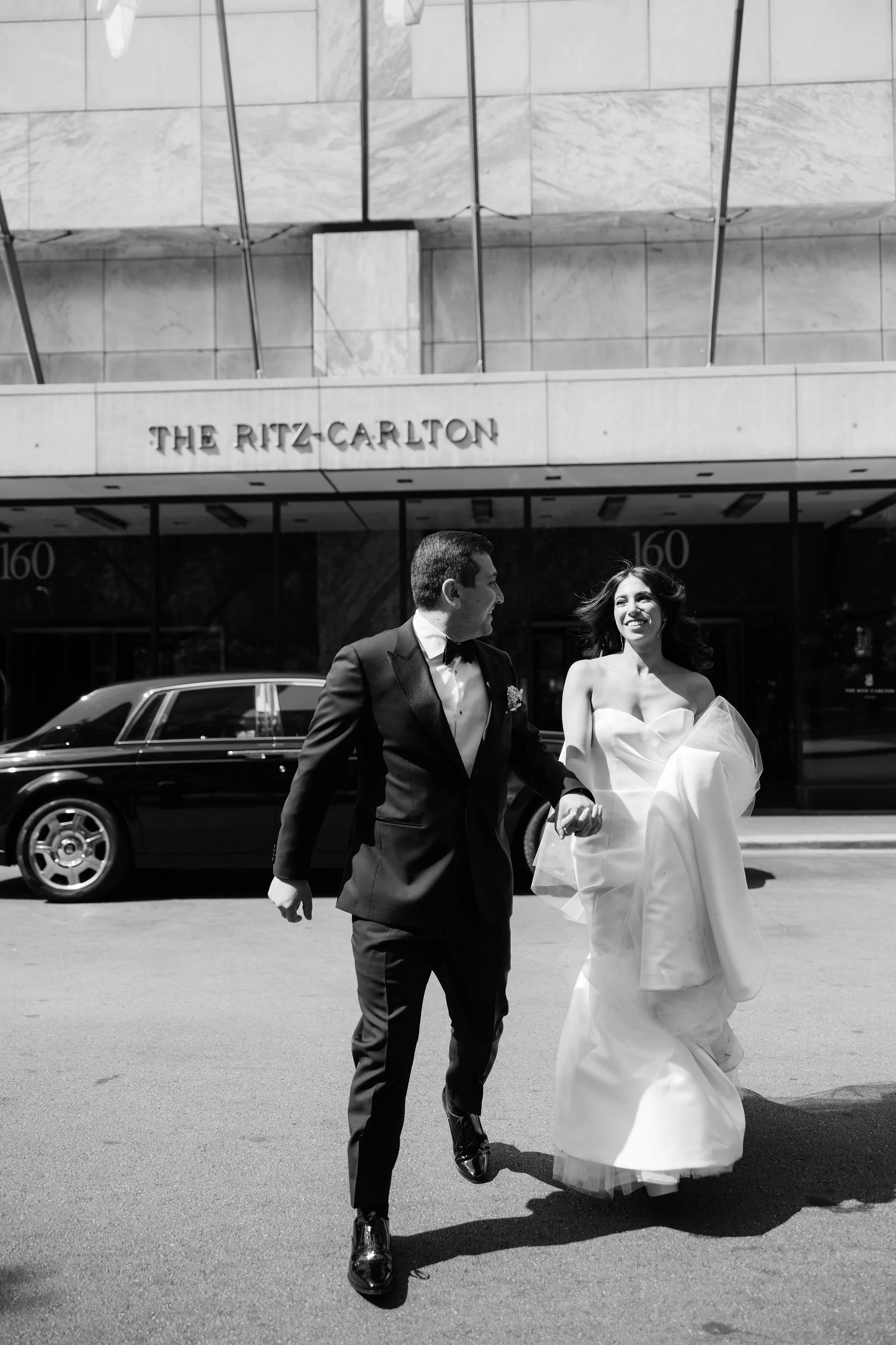 A newlywed couple in wedding attire walking in front of The Ritz-Carlton hotel, with a vintage car parked behind them, black and white photograph.