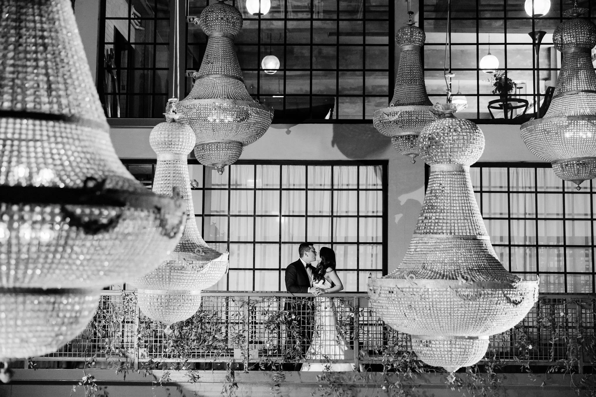 A black and white photo of a couple in formal attire, standing on an indoor balcony, surrounded by large, ornate chandeliers.