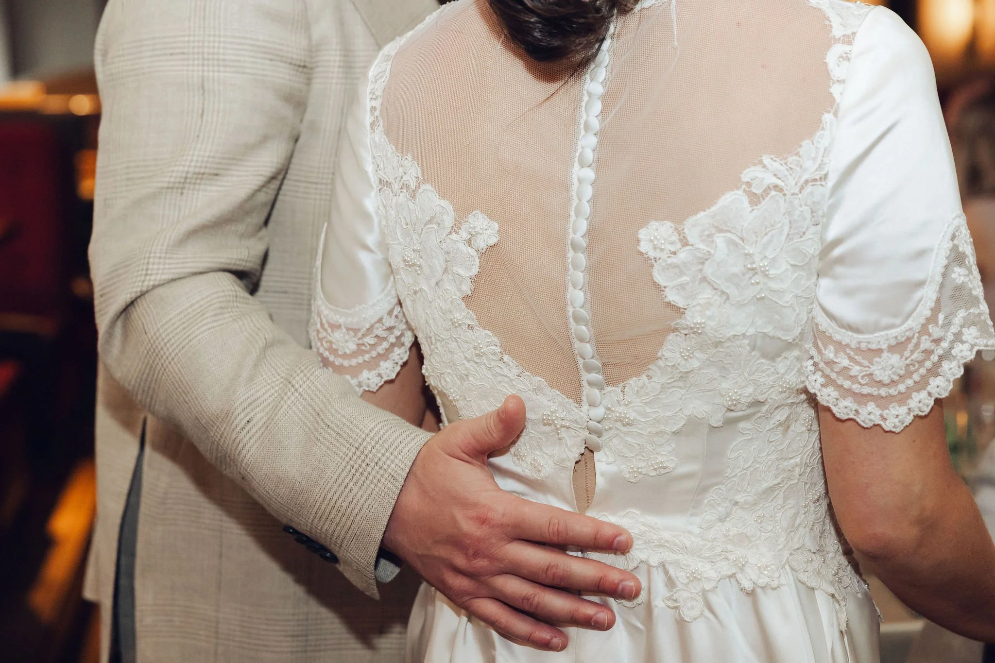 A person wearing a white wedding dress with lace and pearl details is being embraced from behind by someone in a light-colored suit.