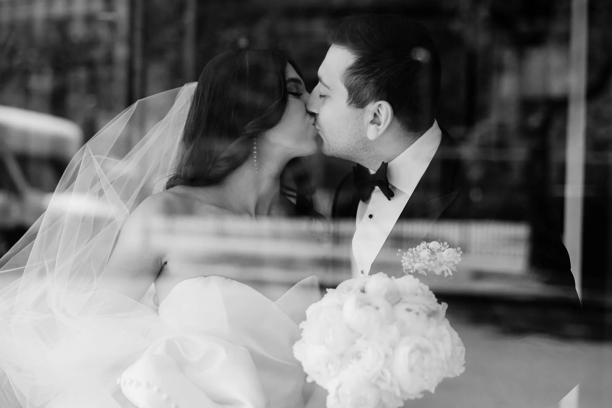 Black and white photo of a bride and groom sharing a kiss, with the bride holding a bouquet of roses, seen through a glass window.