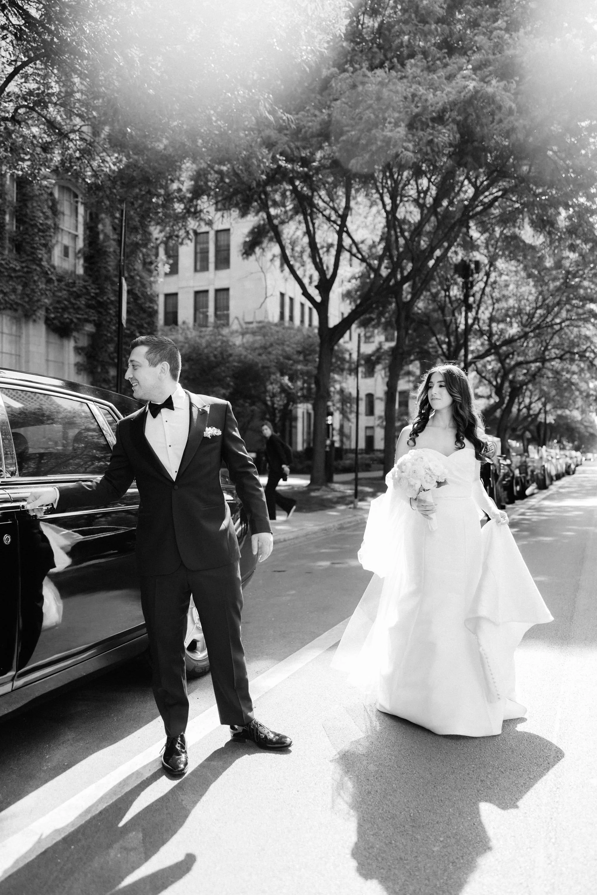 Black and white photo of a bride in a white wedding dress holding a bouquet and a groom in a black tuxedo standing next to a car on a city street lined with trees and parked cars.